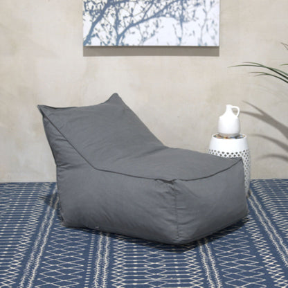 The Ettie Water Resistant Fabric Bean Bag Chair in gray sits on a patterned blue rug beside a white side table with a white pitcher, while wall art and green leaves appear in the background.