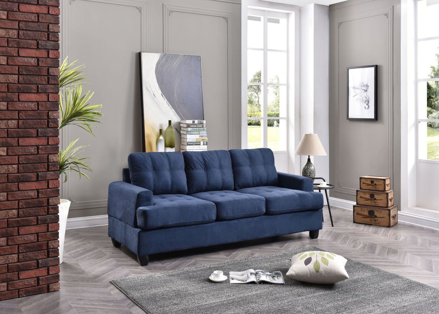 A modern living room featuring the Sandridge sofa in blue, a gray rug, a white pillow with green leaf prints, a potted plant, a lamp on a small wooden chest, stacked books, and abstract art against light gray walls.