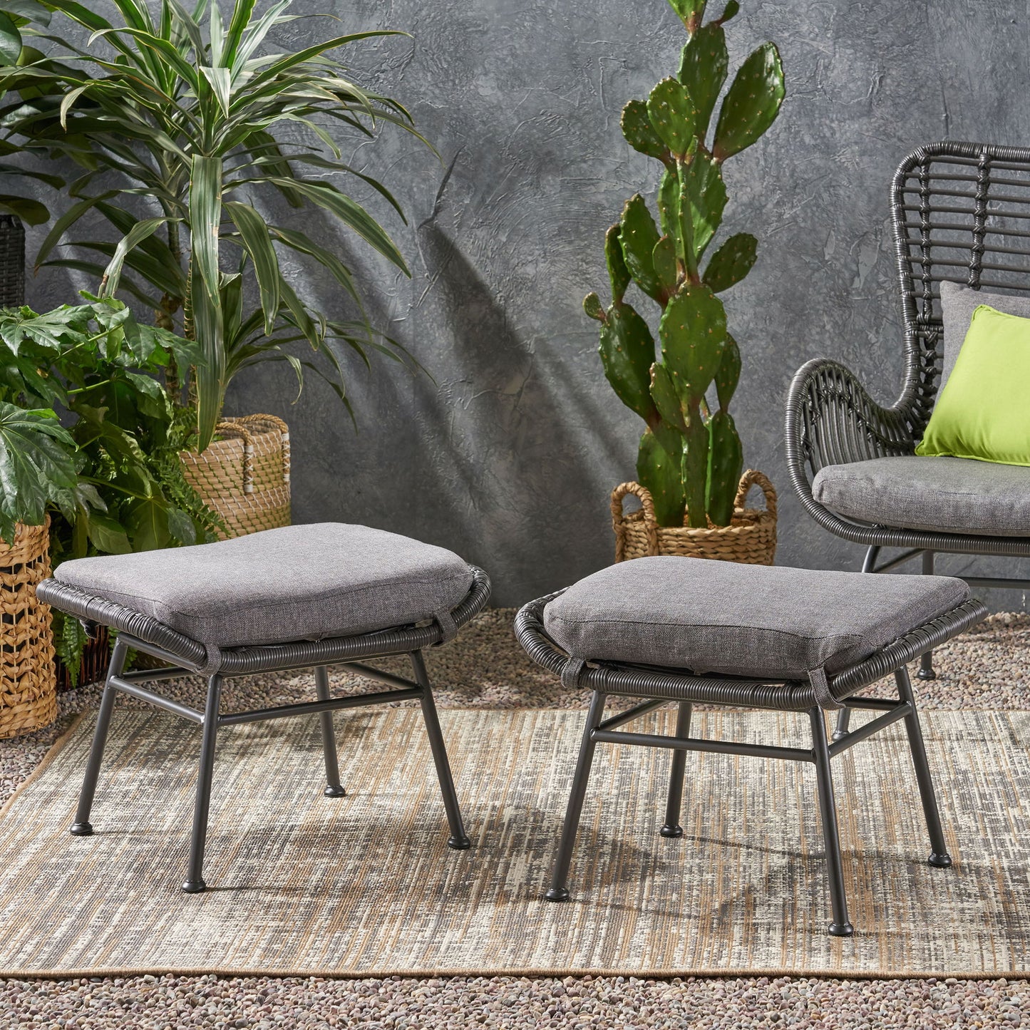 Two Outdoor Modern Boho Wicker Ottomans With Cushions (Set of 2) are placed on a textured rug outdoors, surrounded by potted green plants and a wicker chair with a green cushion against a gray wall.