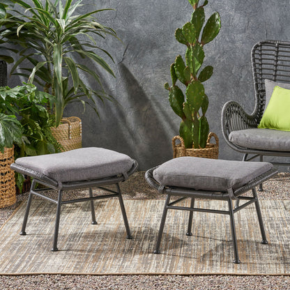 Two Outdoor Modern Boho Wicker Ottomans With Cushions (Set of 2) are placed on a textured rug outdoors, surrounded by potted green plants and a wicker chair with a green cushion against a gray wall.