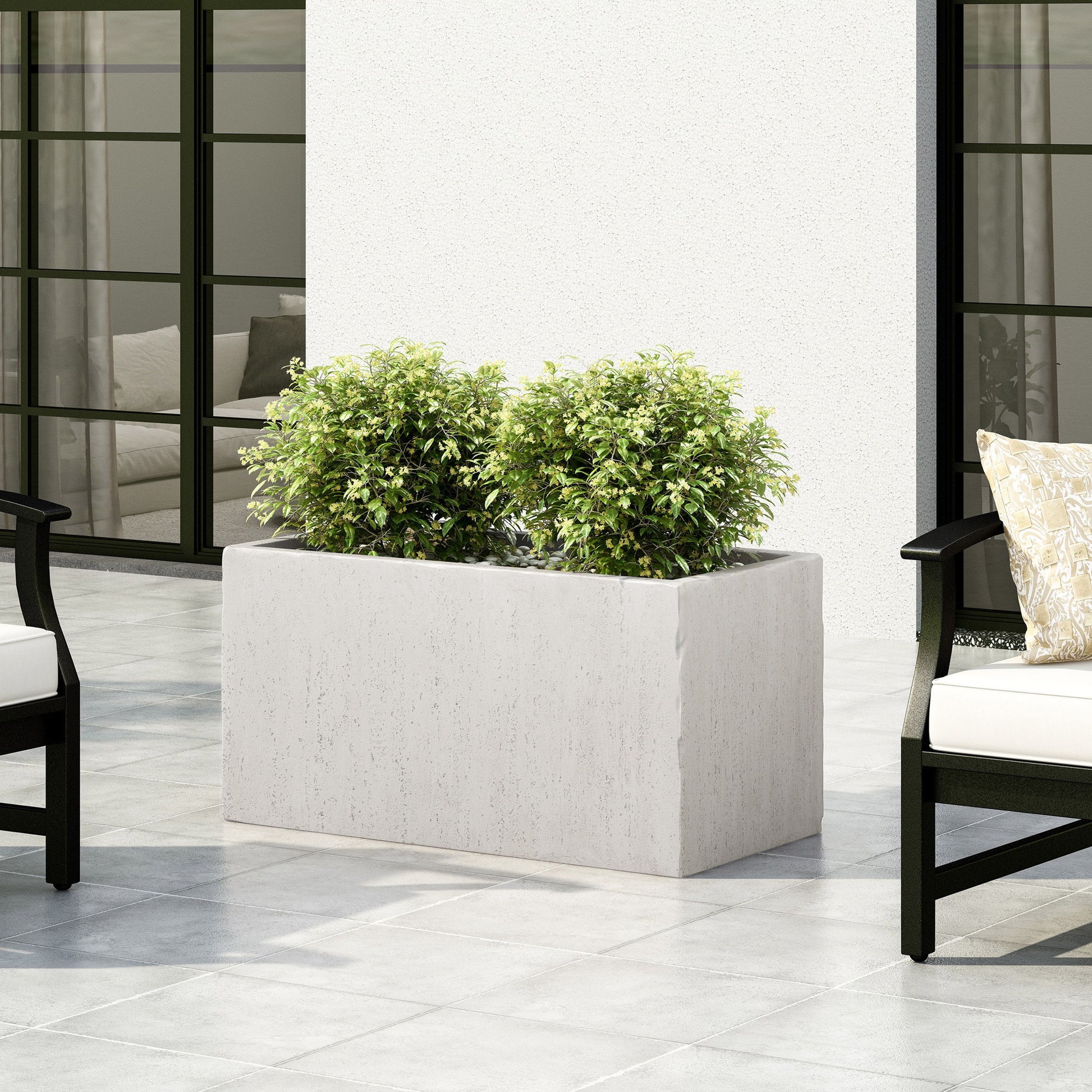A Modern Outdoor Planter with two green leafy bushes is on a tiled patio, surrounded by black outdoor chairs with white cushions and set against a white wall with large windows.