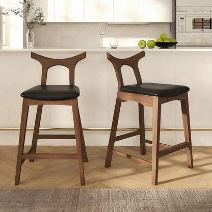 Two Hester Upholstered Square Bar Chairs (Set of 2) with black seats are placed in front of a white kitchen island. The background features white cabinets, a vase with branches, a glass, and a bowl of green apples.