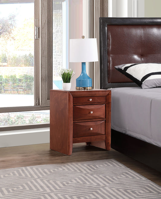 The Marilla Nightstand with three drawers stands beside a bed with a leather headboard, topped with a blue lamp and a small potted plant. A window and geometric-patterned rug complete the scene.