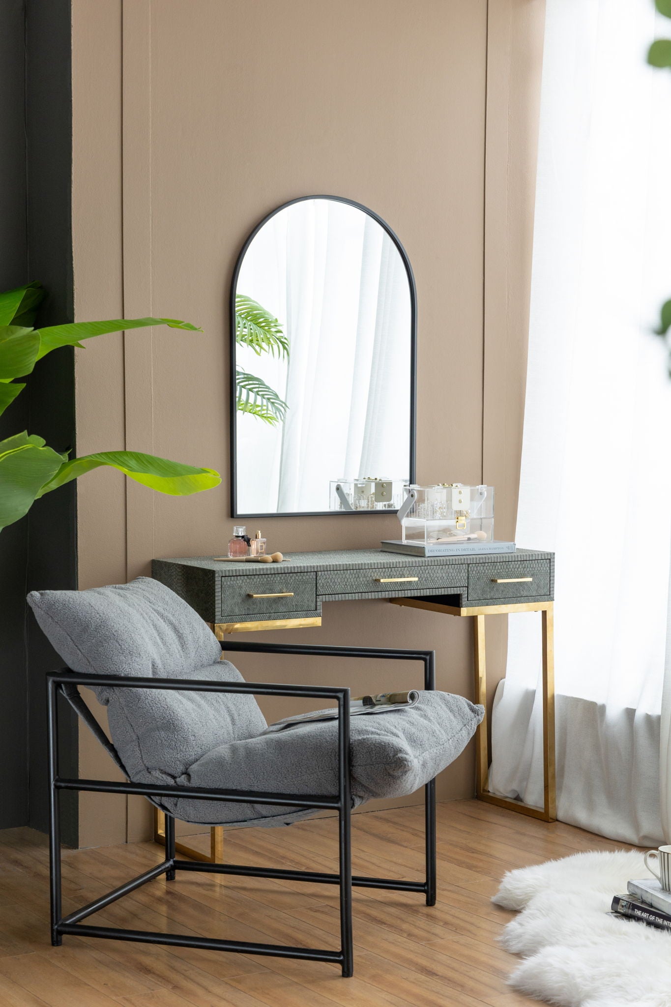 A modern vanity beside a window features the Arched Mirror With Metal Frame, a gold and gray desk, makeup items, sheer curtains, a gray cushioned chair with black metal frame on wood flooring, and a nearby green plant.