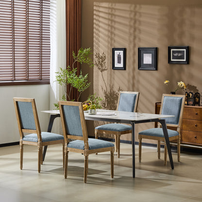 A dining room features a rectangular table, four French Country Style Dining Chairs with upholstered seats and fluted legs, a plant, and three framed pictures on a beige wall. Sunlight streams through window blinds on the left.