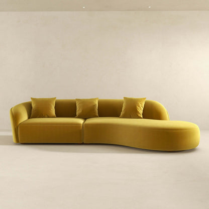 The Elijah - Japandi Style Curvy Sectional Sofa in mustard yellow features four matching square pillows, placed in a minimalistic room with a plain beige wall and light-colored floor.