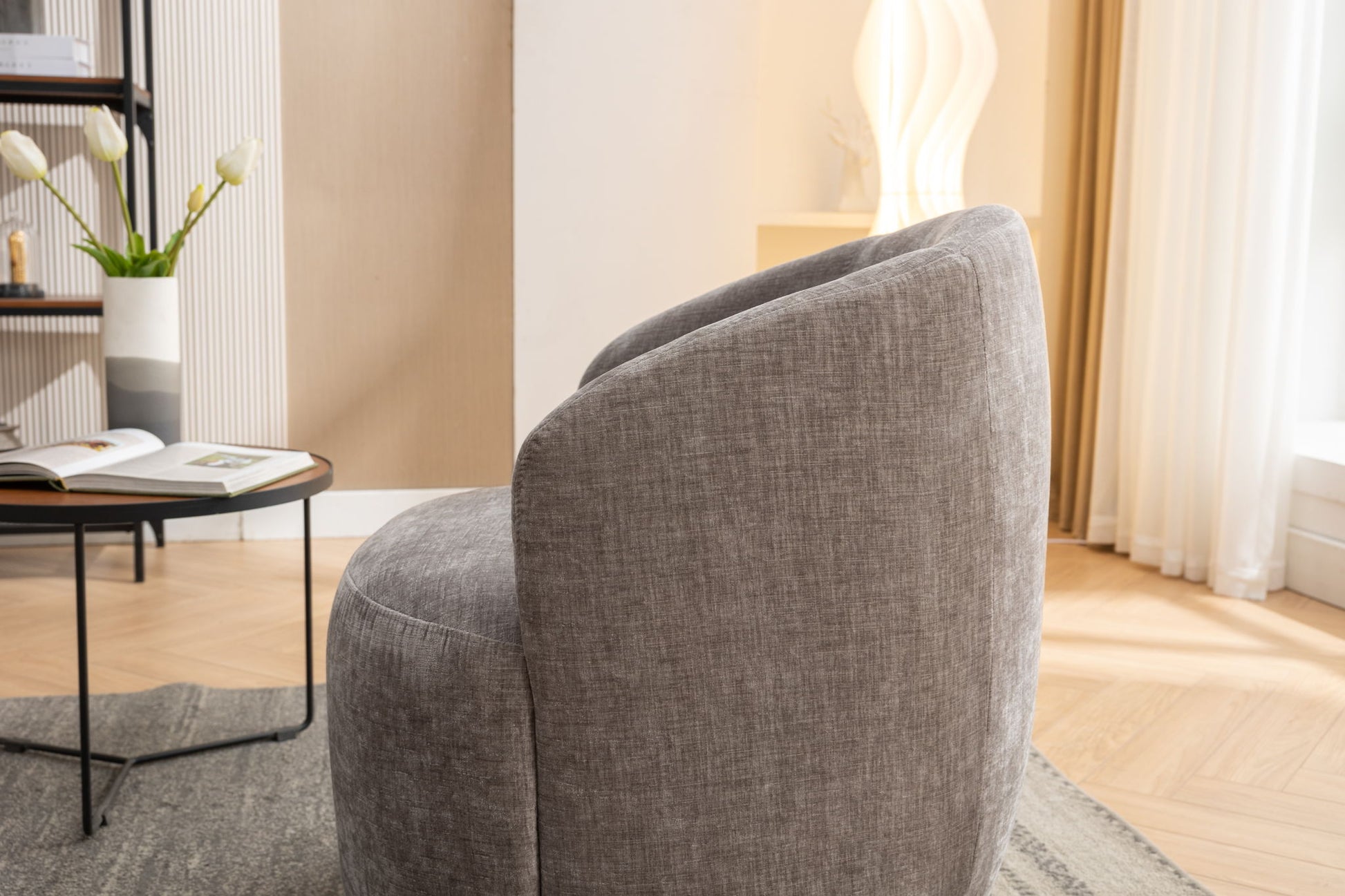 A modern living room features the Chenille Fabric Swivel Accent Armchair Barrel Chair with powder-coated metal ring, a black side table with a book and white flowers, a gray rug, sculptural white lamp, and sheer curtains letting in natural light.