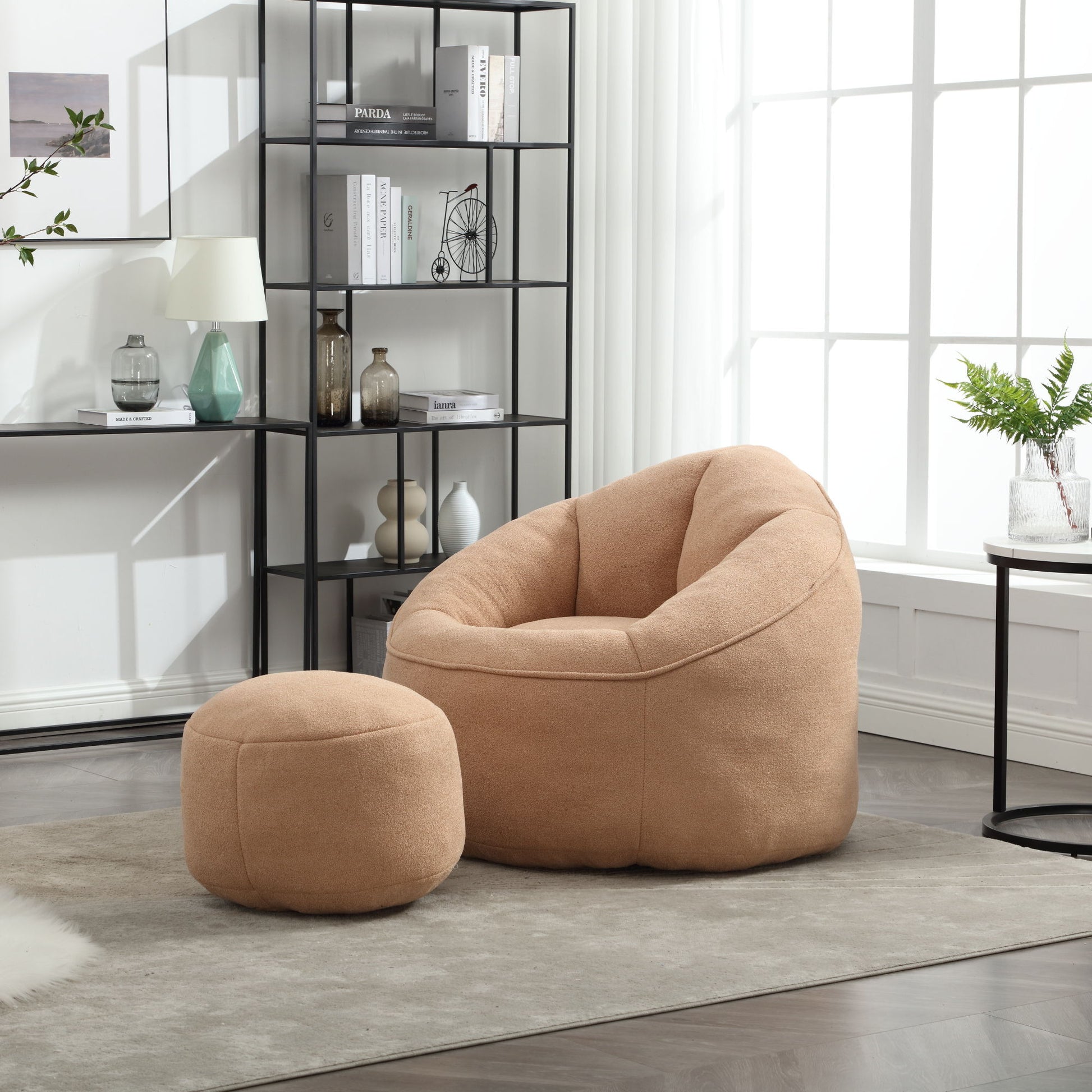 The Bedding Bean Bag Sofa Chair High Pressure Foam with Footrest, in tan, sits on a beige rug in a bright, modern living room. Behind it, a black metal bookshelf displays books and decor beneath a sunlit window.