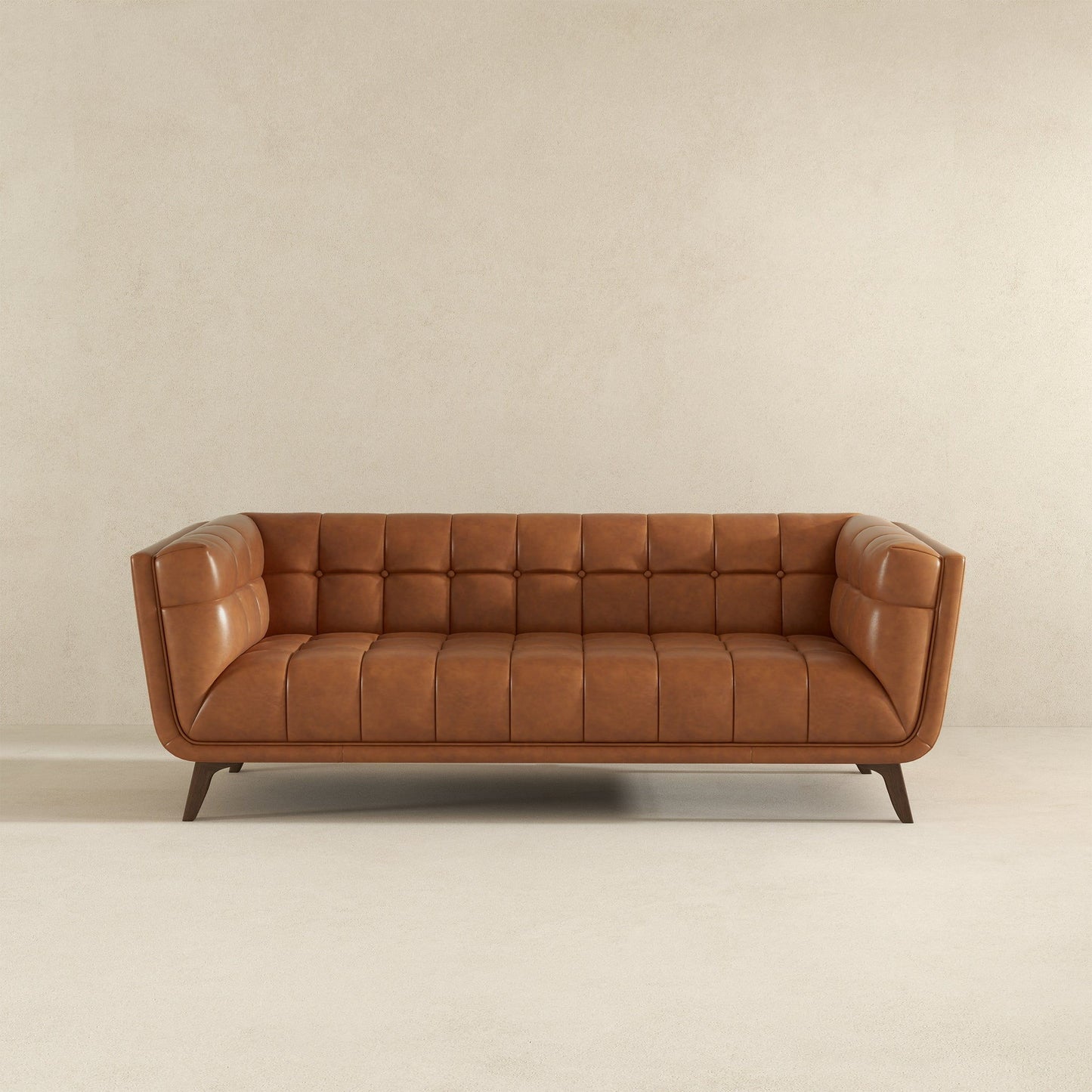 The Addison Mid-Century Modern Design Tufted Sofa in tan leather, featuring tufted cushions and angled wooden legs, is placed against a plain beige wall on a light carpeted floor.