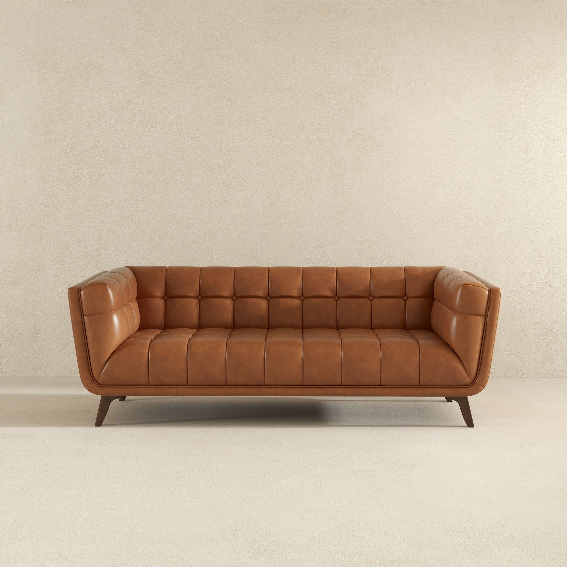 The Addison Mid-Century Modern Design Tufted Sofa in tan leather, featuring tufted cushions and angled wooden legs, is placed against a plain beige wall on a light carpeted floor.