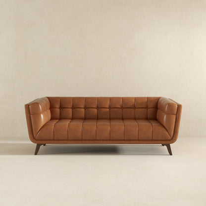 The Addison Mid-Century Modern Design Tufted Sofa in tan leather, featuring tufted cushions and angled wooden legs, is placed against a plain beige wall on a light carpeted floor.