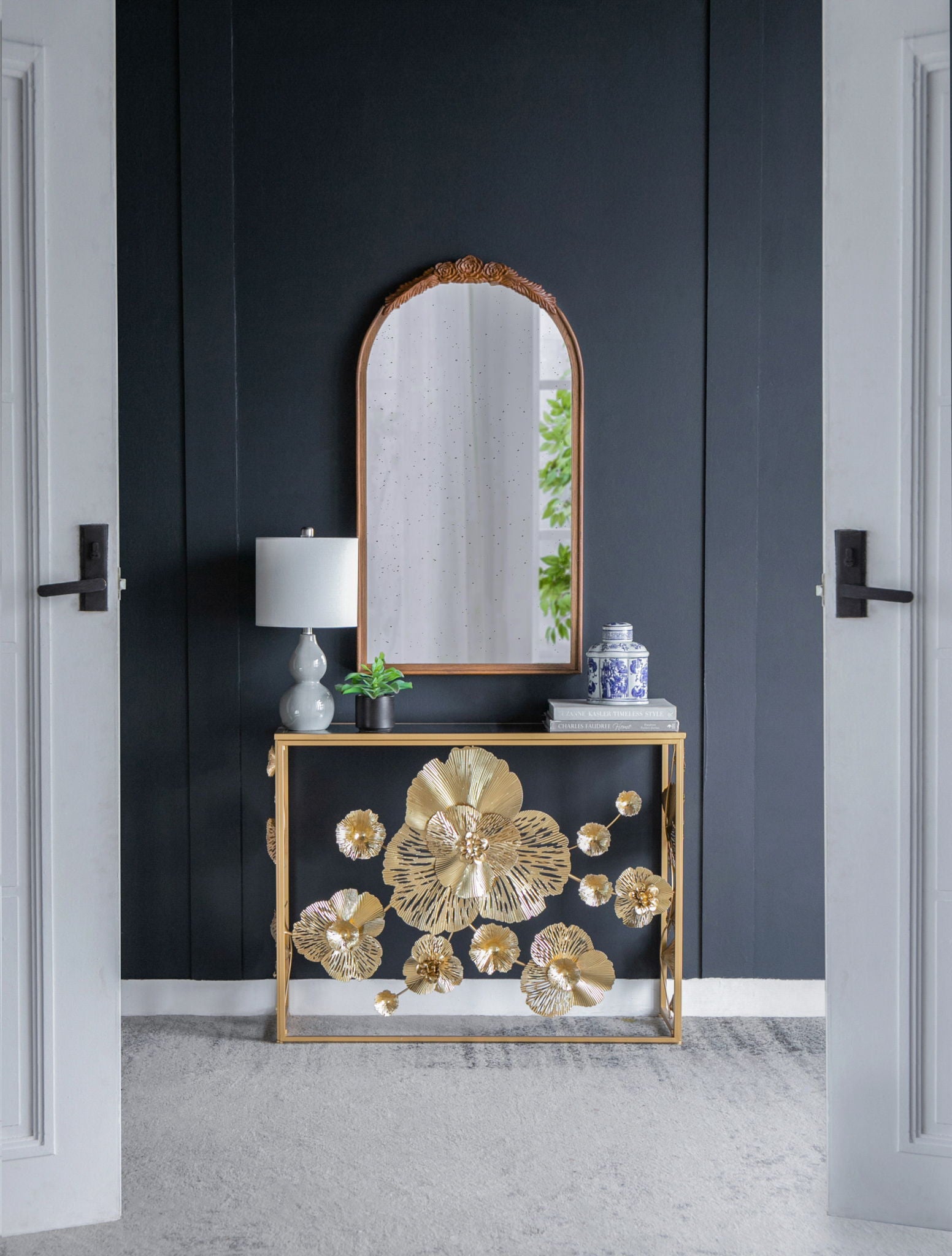 A modern entryway featuring a black wall, a gold Console Table With Mirrored Glass Top, a table lamp, blue-and-white vase, and an arched mirror reflecting greenery.