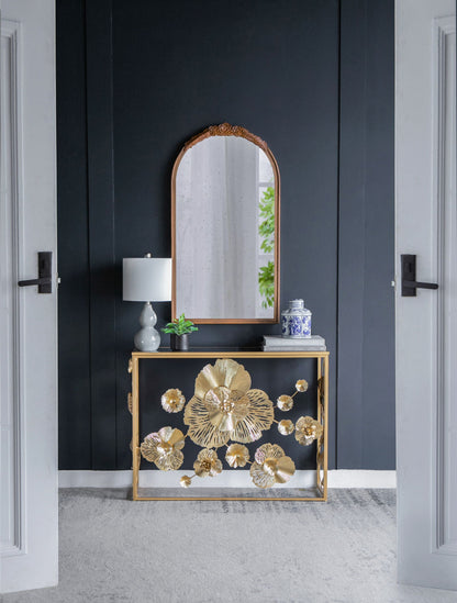 A modern entryway featuring a black wall, a gold Console Table With Mirrored Glass Top, a table lamp, blue-and-white vase, and an arched mirror reflecting greenery.