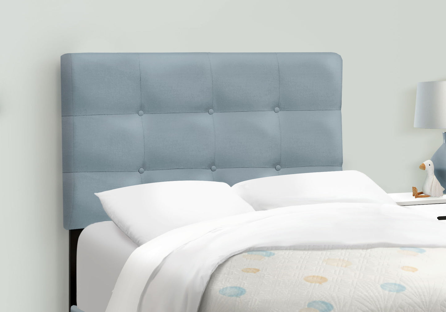 A neatly made bed with white bedding, a transitional upholstered bed frame in light blue, and a nightstand with a white lamp and decorative duck figurine. The quilt features a subtle polka dot pattern.
