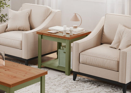 Two beige armchairs with pillows sit beside the Couch.com Vineyard Side Table in sage green and fruitwood, topped with books, candles, and a lamp. A matching coffee table and a textured rug complete the cozy, light-filled room.