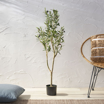 An Artificial Olive Tree with slender branches and green leaves stands on a light wood floor beside a woven chair and blue cushion, set against a textured white wall.