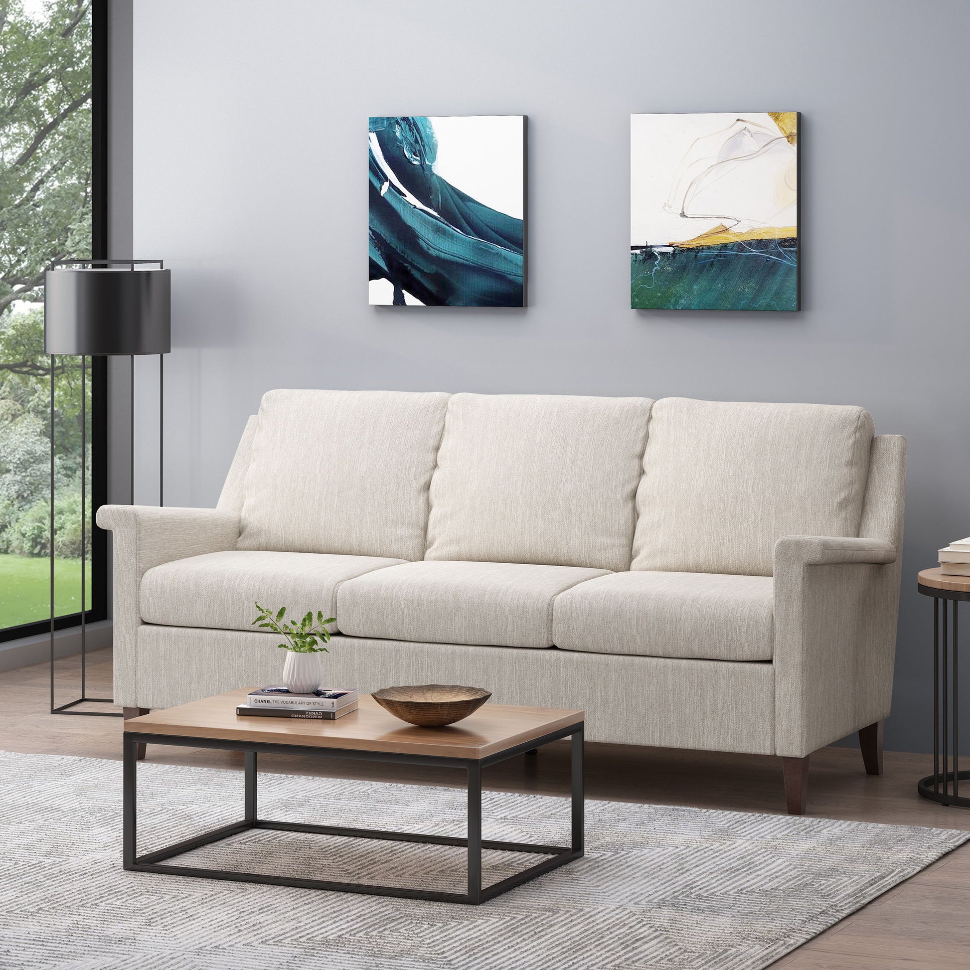 A modern living room features a 3 Seater Contemporary Design Sofa in beige, a wooden coffee table with a book and bowl, floor lamp, light gray rug, two abstract paintings on a gray wall, and a window overlooking greenery.