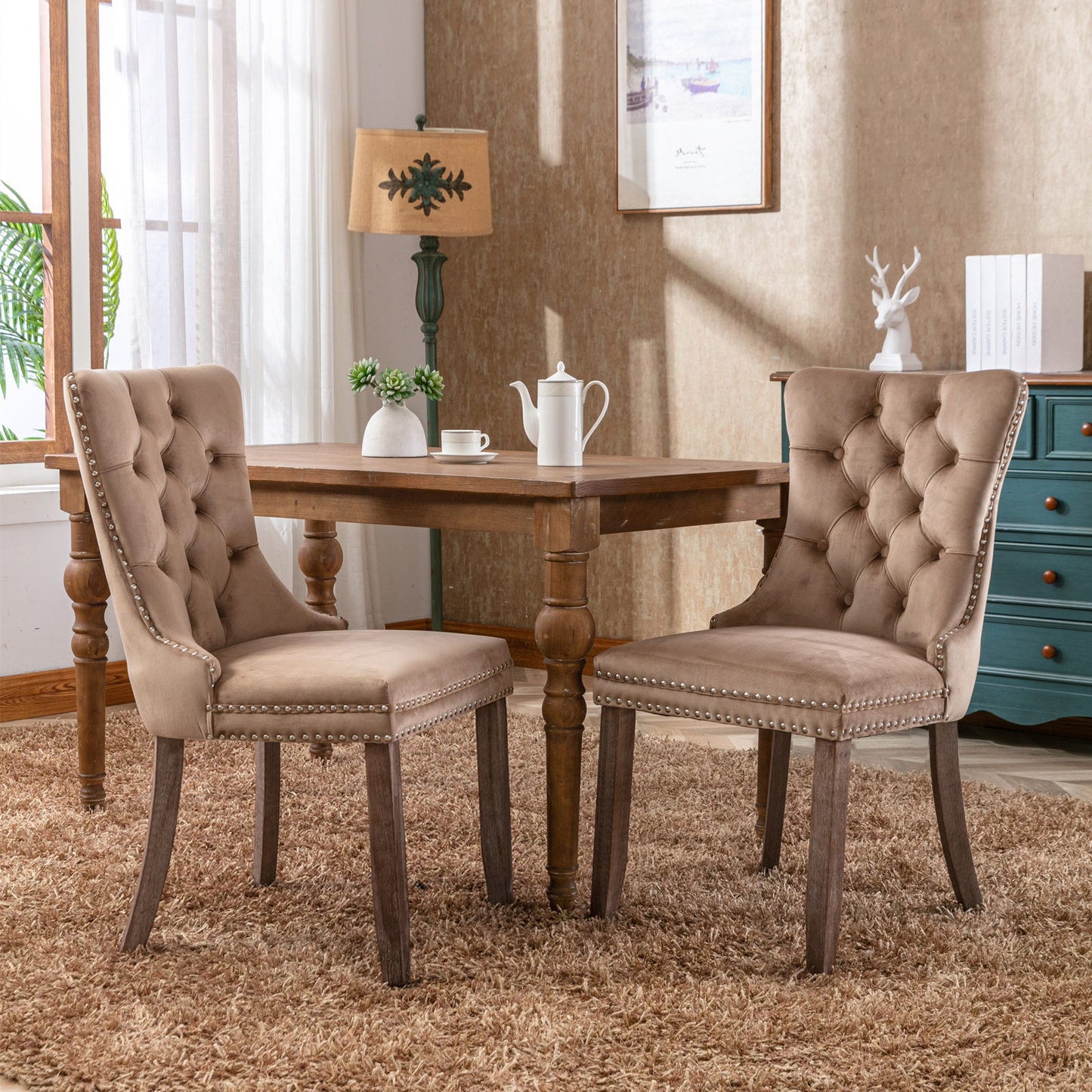 Two Nikki Modern Velvet Tufted Dining Chairs with nailhead trim sit at a wooden square table on a brown rug. A teapot and cups rest on the table, with a window, lamp, artwork, and blue dresser visible in the background.