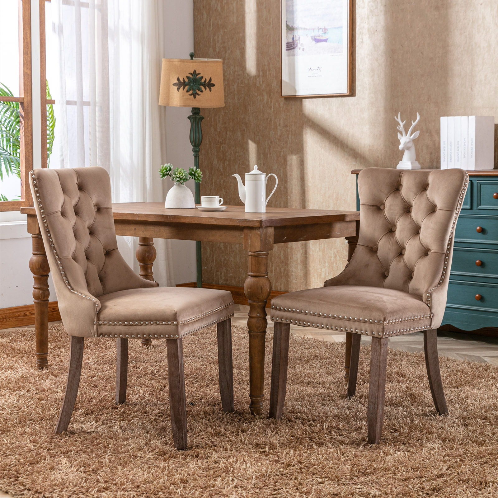Two Nikki Modern Velvet Tufted Dining Chairs with nailhead trim sit at a wooden square table on a brown rug. A teapot and cups rest on the table, with a window, lamp, artwork, and blue dresser visible in the background.