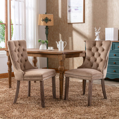Two Nikki Modern Velvet Tufted Dining Chairs with nailhead trim sit at a wooden square table on a brown rug. A teapot and cups rest on the table, with a window, lamp, artwork, and blue dresser visible in the background.