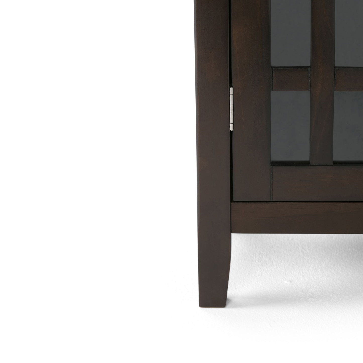 Close-up of the lower right corner of the Bedford - Storage Media Cabinet, featuring a dark wood finish, glass panel door, silver hinge, and a visible leg on a white floor.
