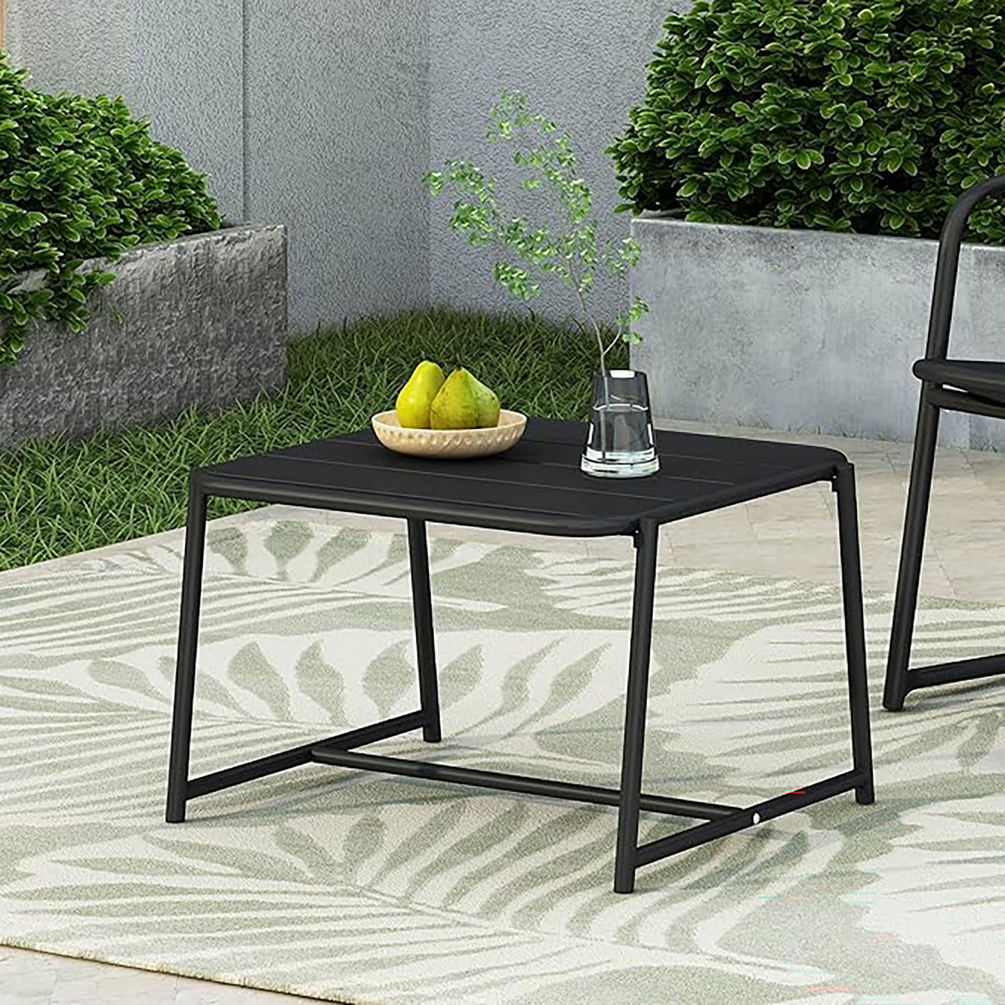 The Conrad - Side Table Outdoor, in black, sits on a patterned rug holding a bowl of pears and a glass of water. Green bushes and gray concrete planters complete the scene.