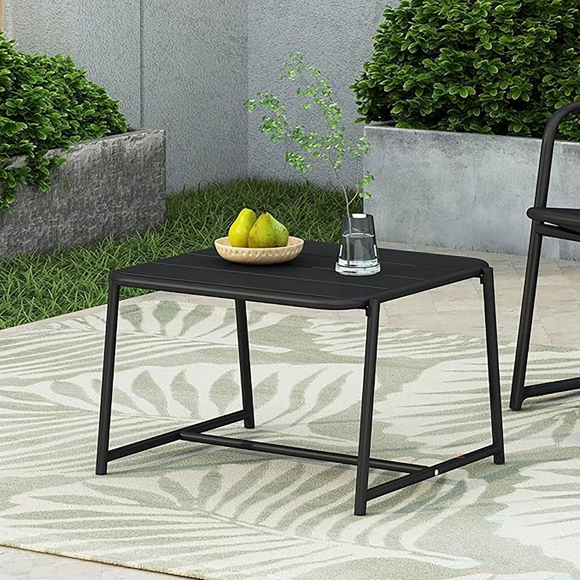 The Conrad - Side Table Outdoor, in black, sits on a patterned rug holding a bowl of pears and a glass of water. Green bushes and gray concrete planters complete the scene.