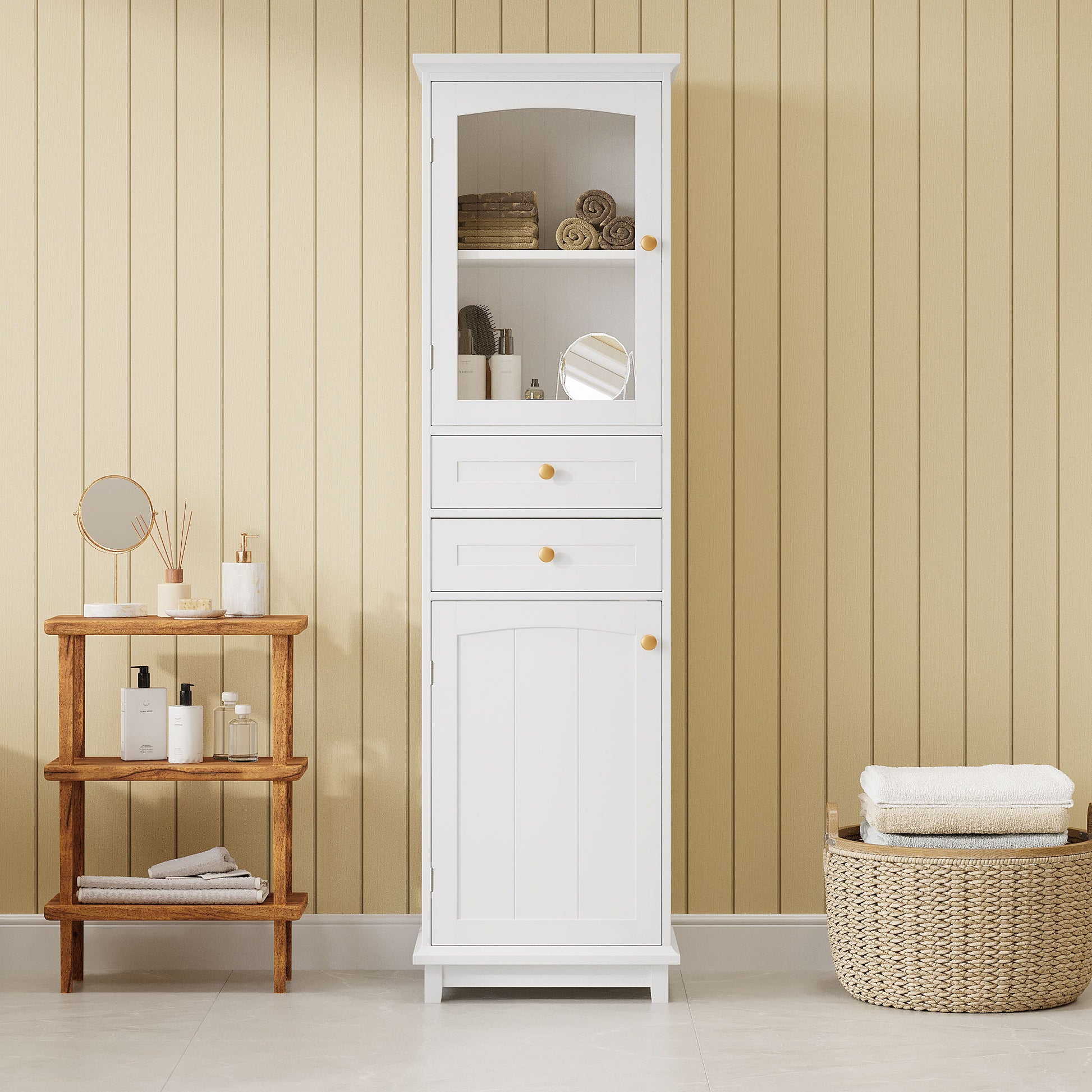 A Tall Bathroom Storage Cabinet With Glass Doors, free-standing with two drawers and adjustable shelves, made of painted MDF board for displaying your favorite items, stands against a beige paneled wall on a light tiled floor.