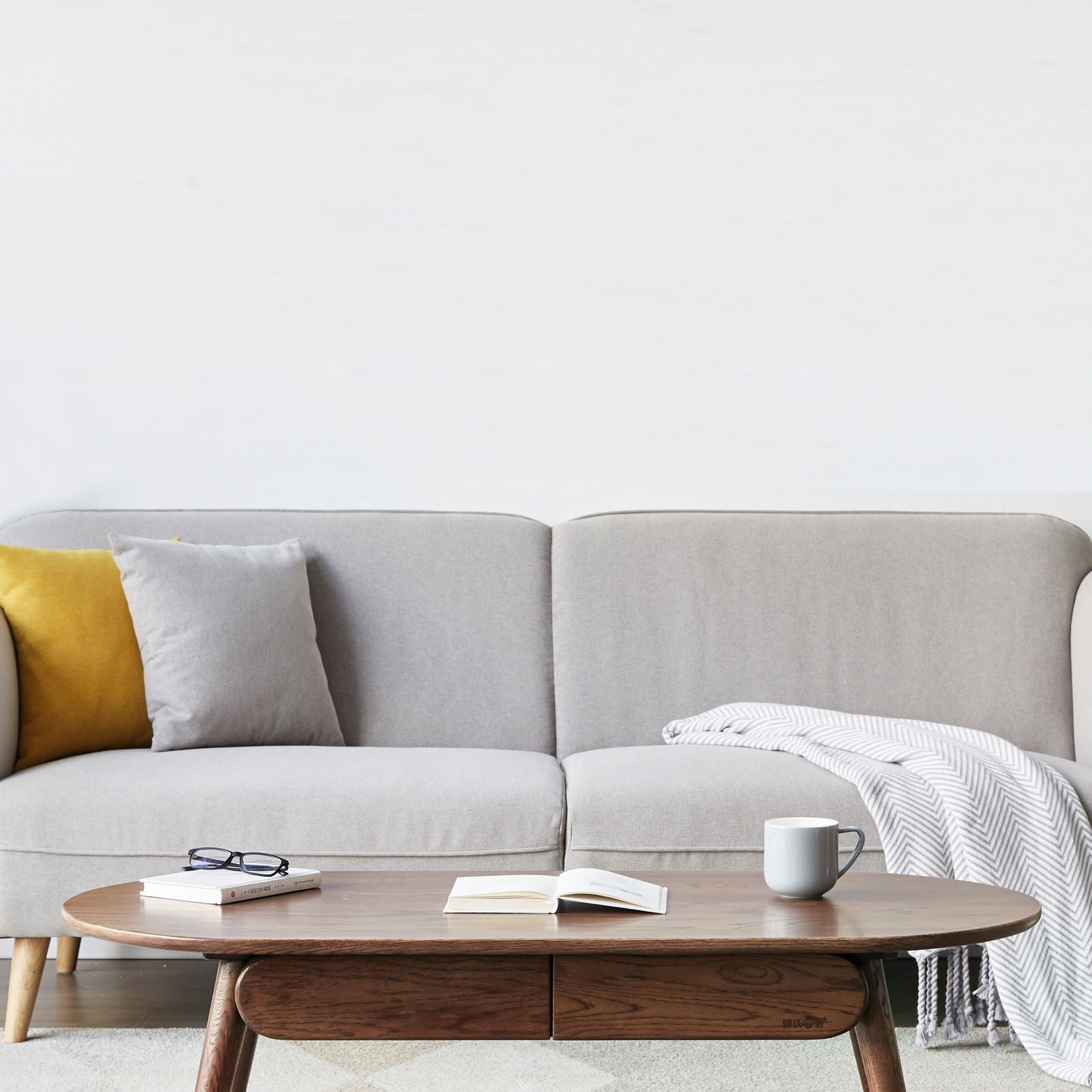 A modern living room with a light gray sofa, yellow and gray cushions, a striped blanket, and the Capsule Centre Low Table With Drawers Coffee Table holding a mug, open book, and eyeglasses against a plain white wall.
