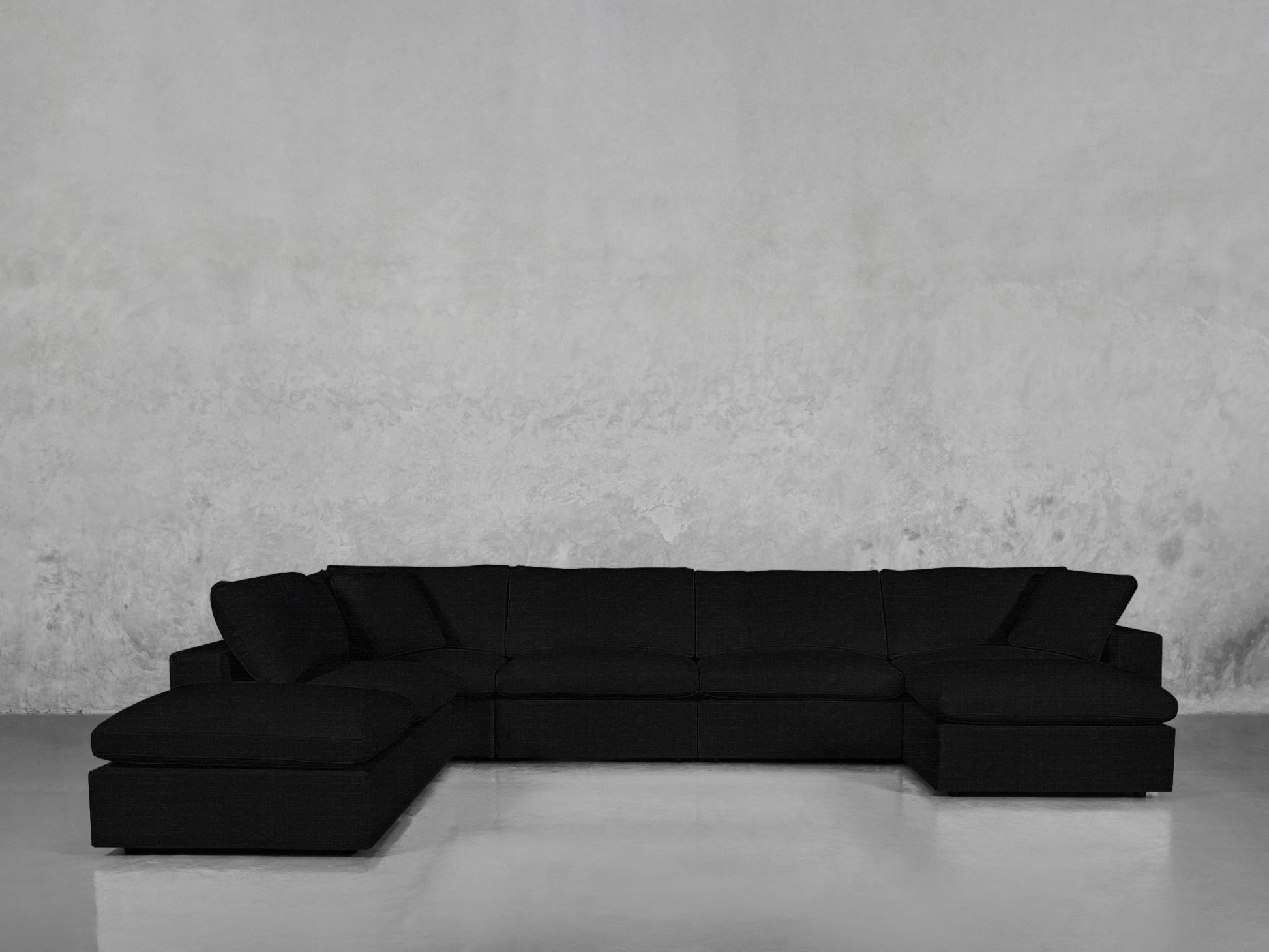 The 7-Seat Modular Chaise Corner Lounger Sectional, a large black modern sofa with deep cushions, is set against a plain textured gray wall on a smooth reflective floor.