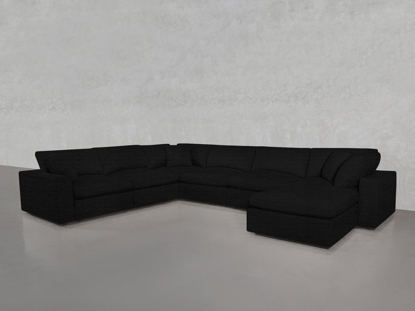 A 7-Seat Modular Chaise Corner Sectional in modern black is arranged in an L-shape within a minimal, light gray room featuring smooth floors and textured walls.