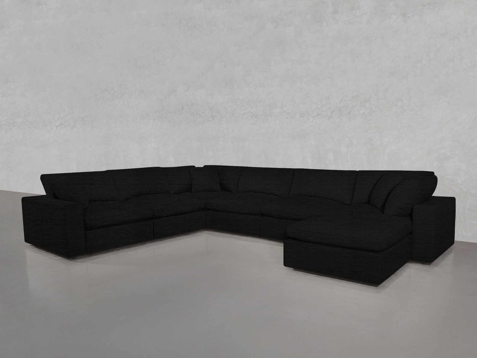 A 7-Seat Modular Chaise Corner Sectional in modern black is arranged in an L-shape within a minimal, light gray room featuring smooth floors and textured walls.