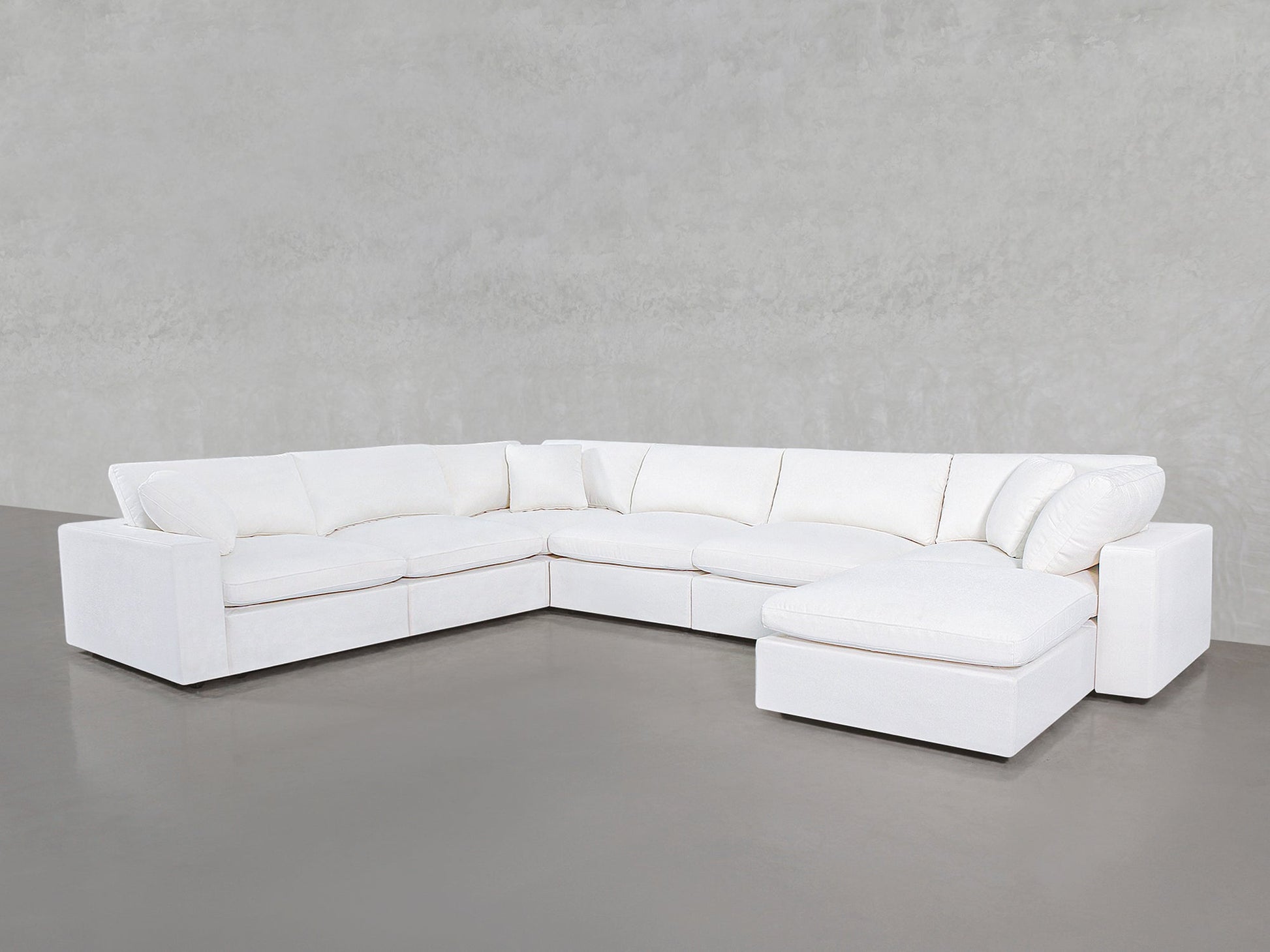 The 7-Seat Modular Chaise Corner Sectional, with plush cushions and a modern white design, sits in the corner of a minimalist room featuring gray concrete floors and a light gray textured wall.