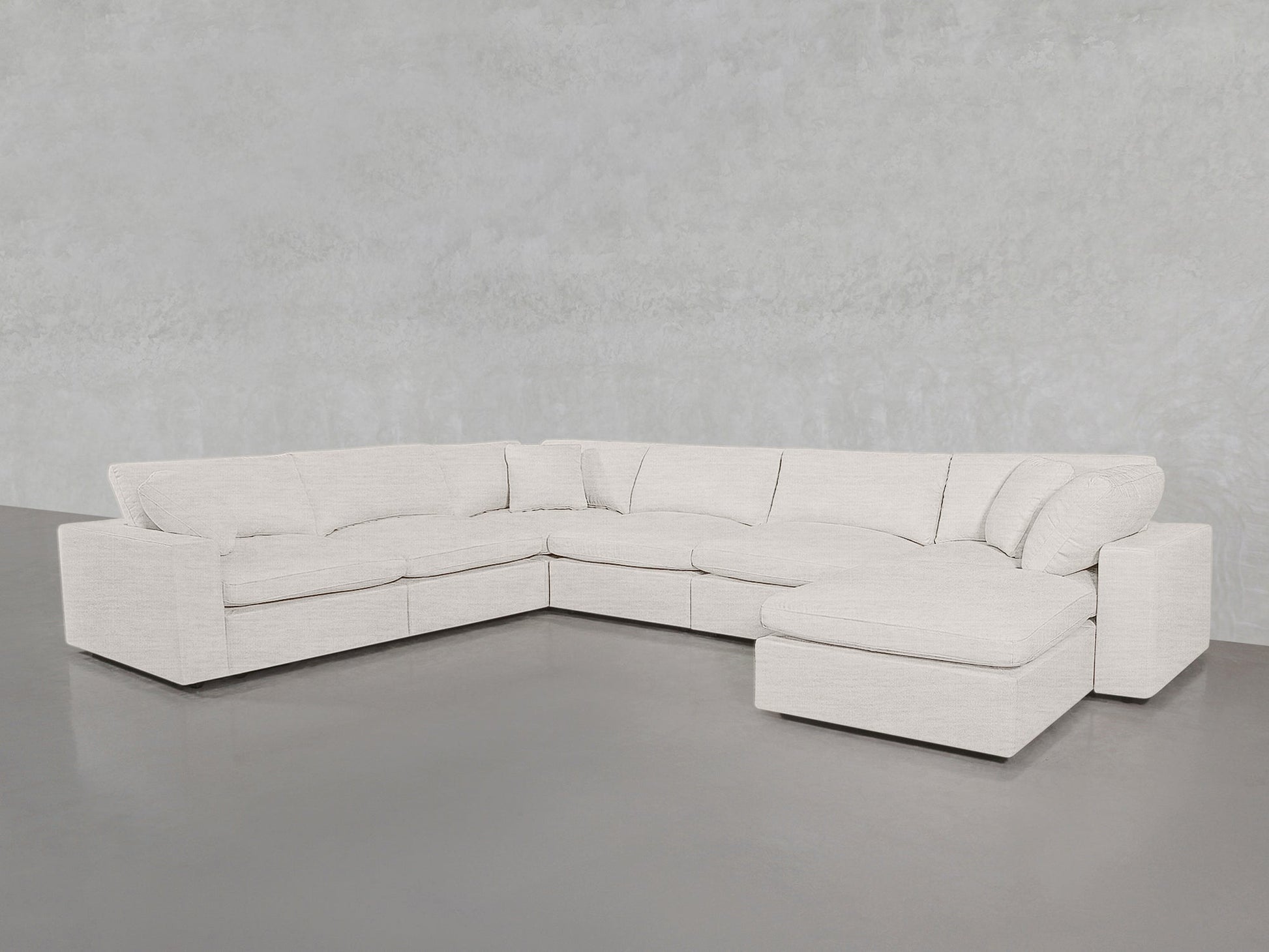 A 7-Seat Modular Chaise Corner Sectional in light beige, featuring plush cushions, is arranged in an L-shape against a minimalist light gray wall and smooth gray floor.