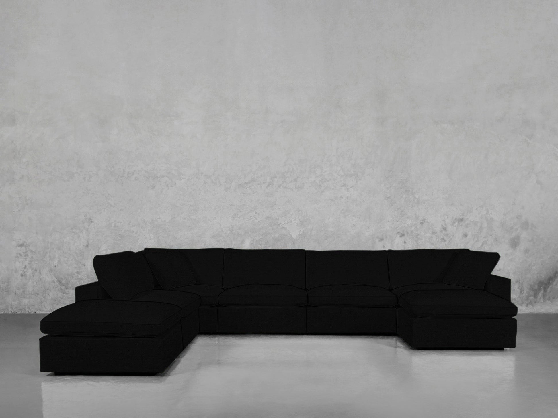 A spacious black 7-Seat Modular Chaise Corner Lounger Sectional with a left-side chaise rests against a plain, light gray concrete wall and a smooth, reflective floor.