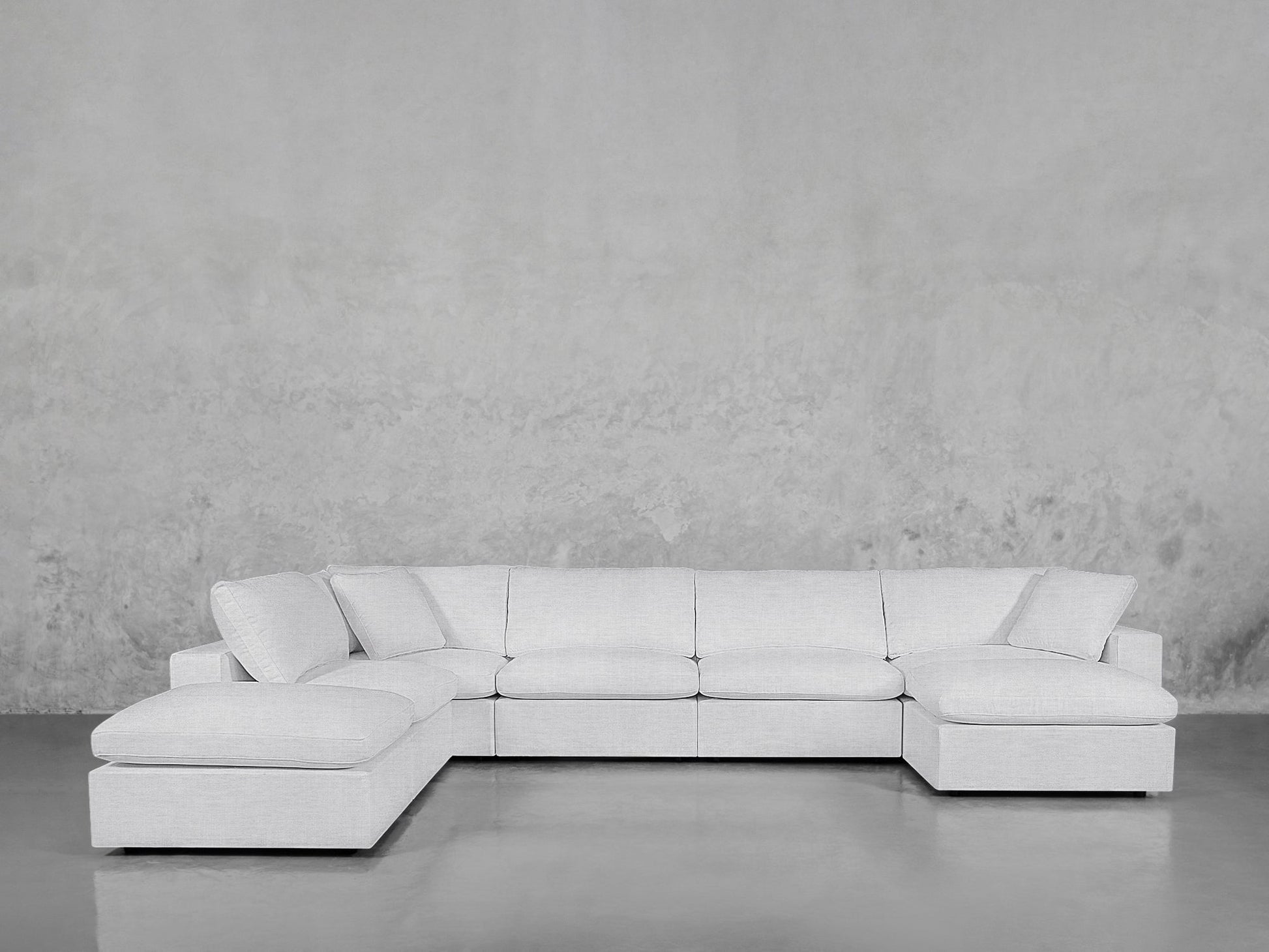 The 7-Seat Modular Chaise Corner Lounger Sectional in light gray, with plush seat and back cushions, wide armrests, and two corner pieces, sits against a textured gray wall on a polished concrete floor.