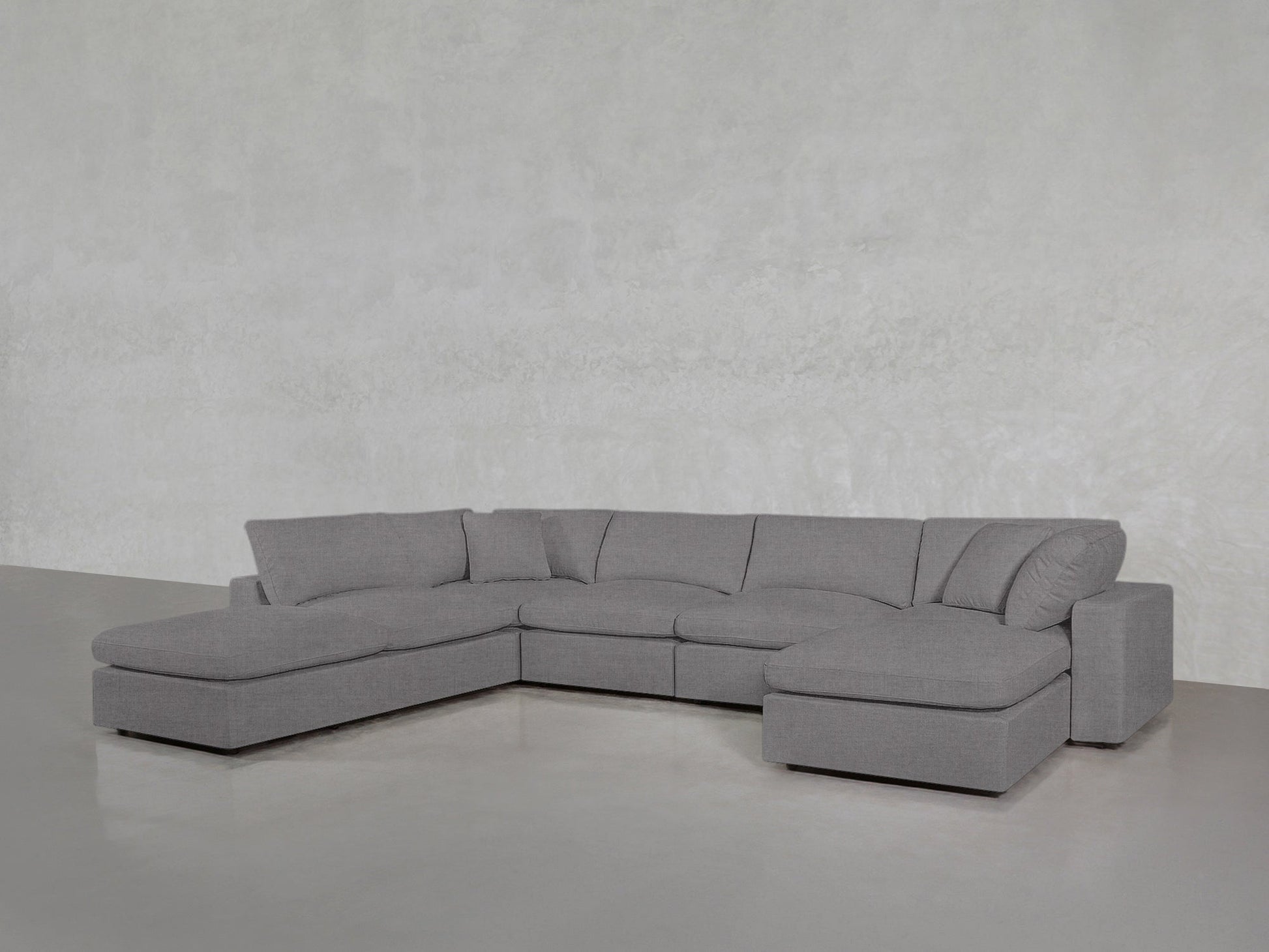 A 7-Seat Modular Chaise Corner Lounger Sectional with plush cushions and clean lines sits on a smooth cement floor against a light gray textured wall.