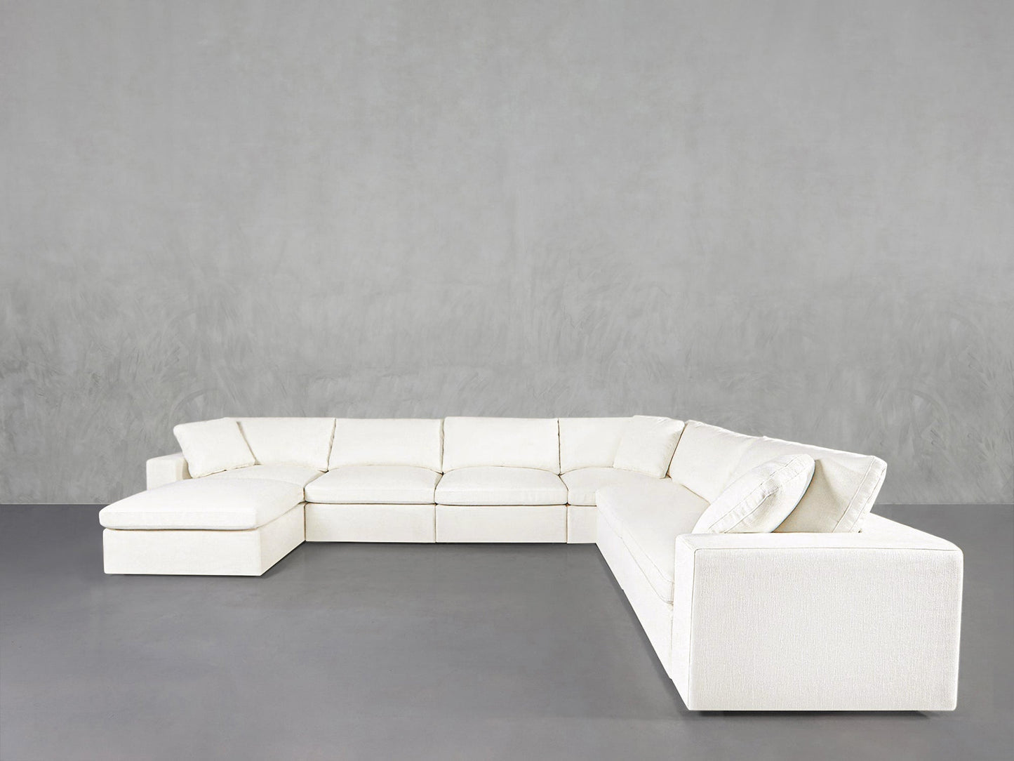 The 7-Seat Modular Chaise Corner Sectional features a large, modern white design with clean lines and a left-side chaise, set against a plain light gray wall and gray floor.