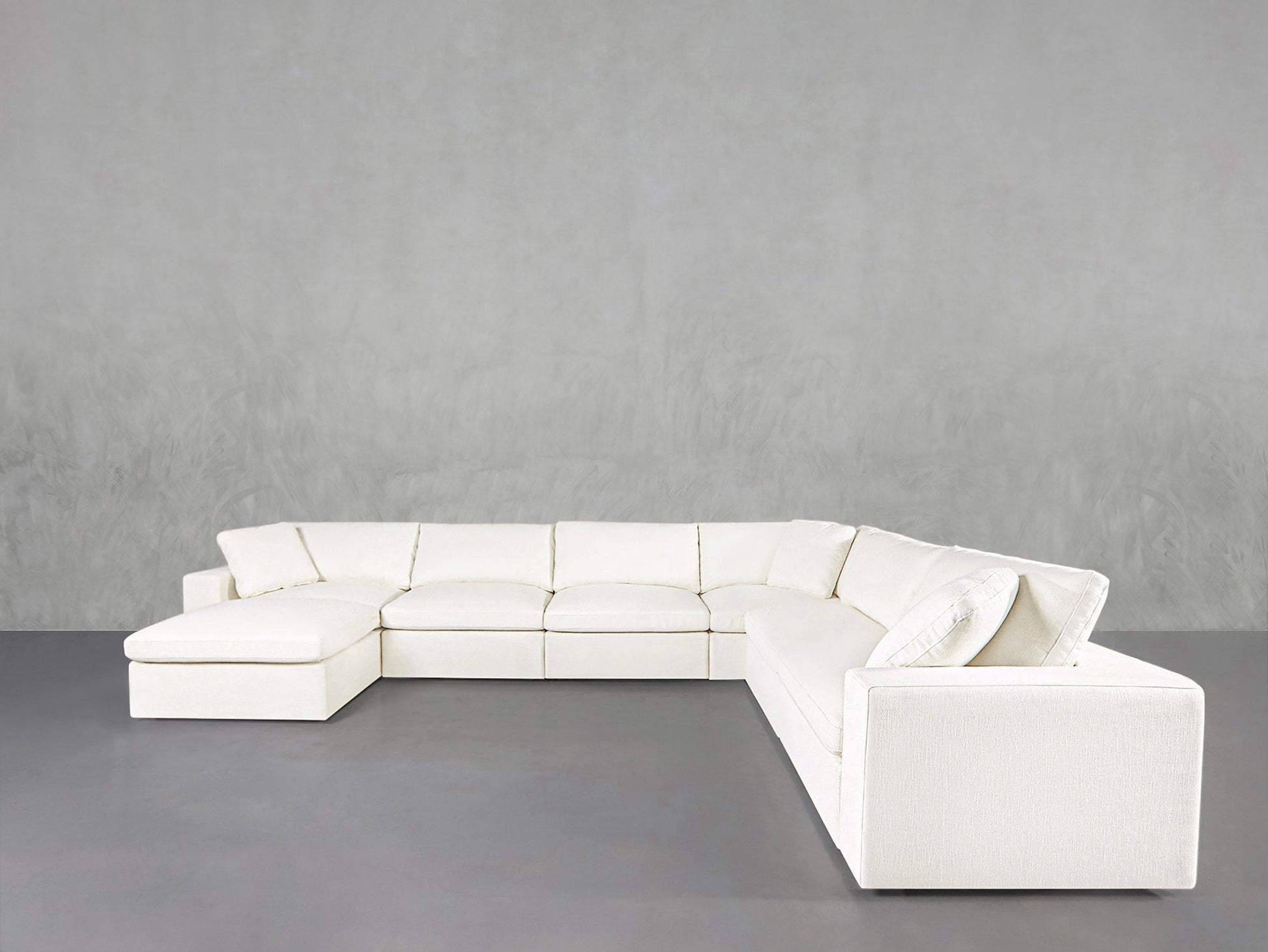 The 7-Seat Modular Chaise Corner Sectional features a large, modern white design with clean lines and a left-side chaise, set against a plain light gray wall and gray floor.