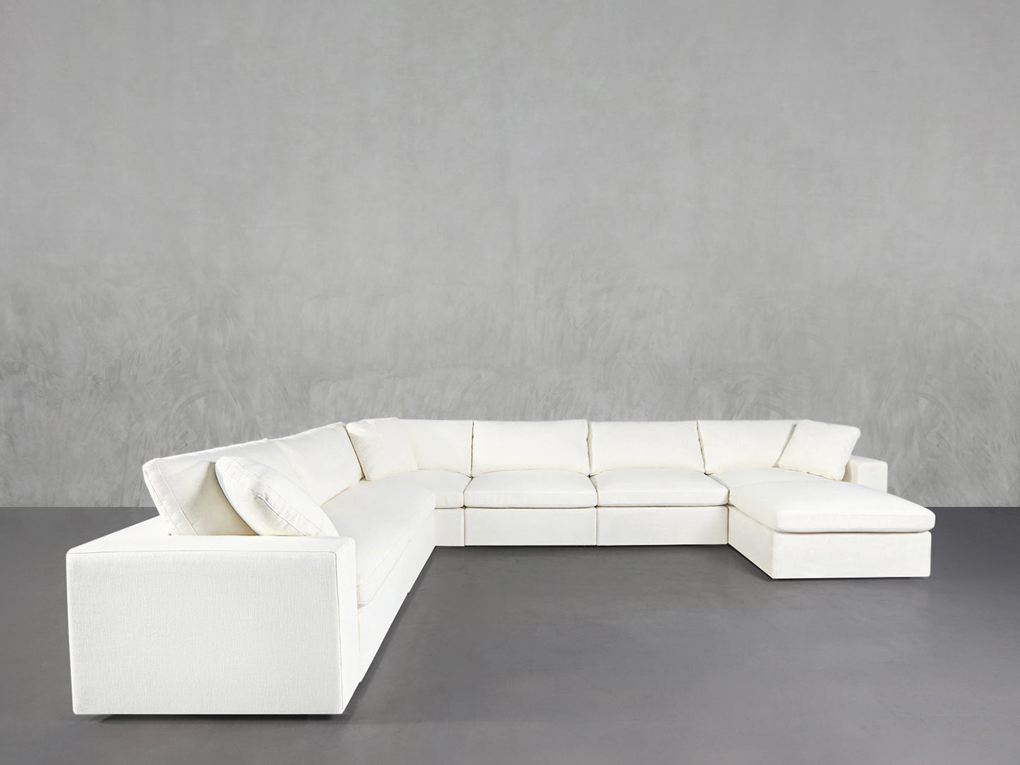 The 7-Seat Modular Chaise Corner Sectional features a large, modern white design with clean lines, placed in a minimalist room with gray flooring and a textured light gray wall behind it.