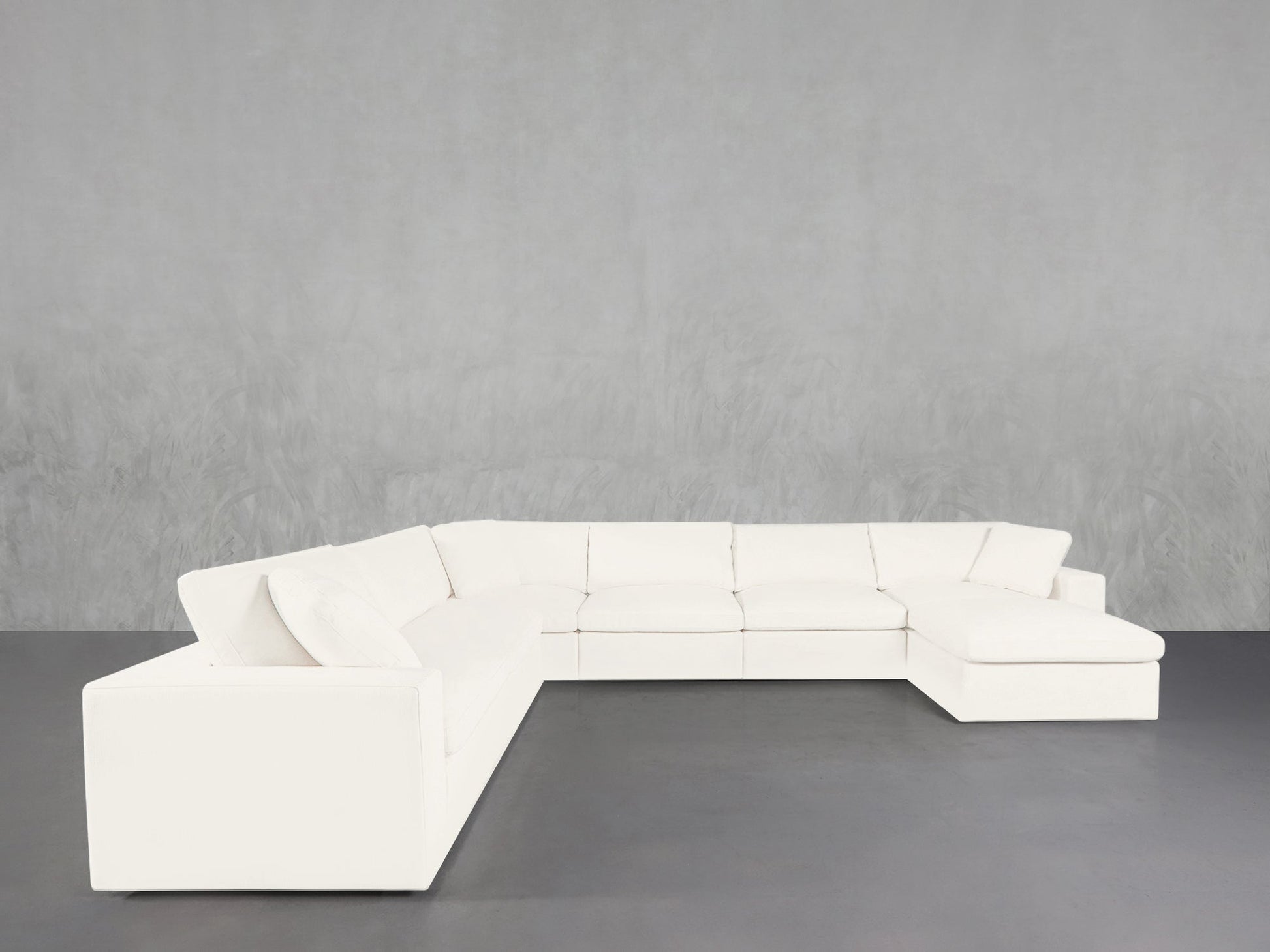 The 7-Seat Modular Chaise Corner Sectional in white, featuring clean lines and a modern design, is set against a plain light gray textured wall and a dark gray floor.