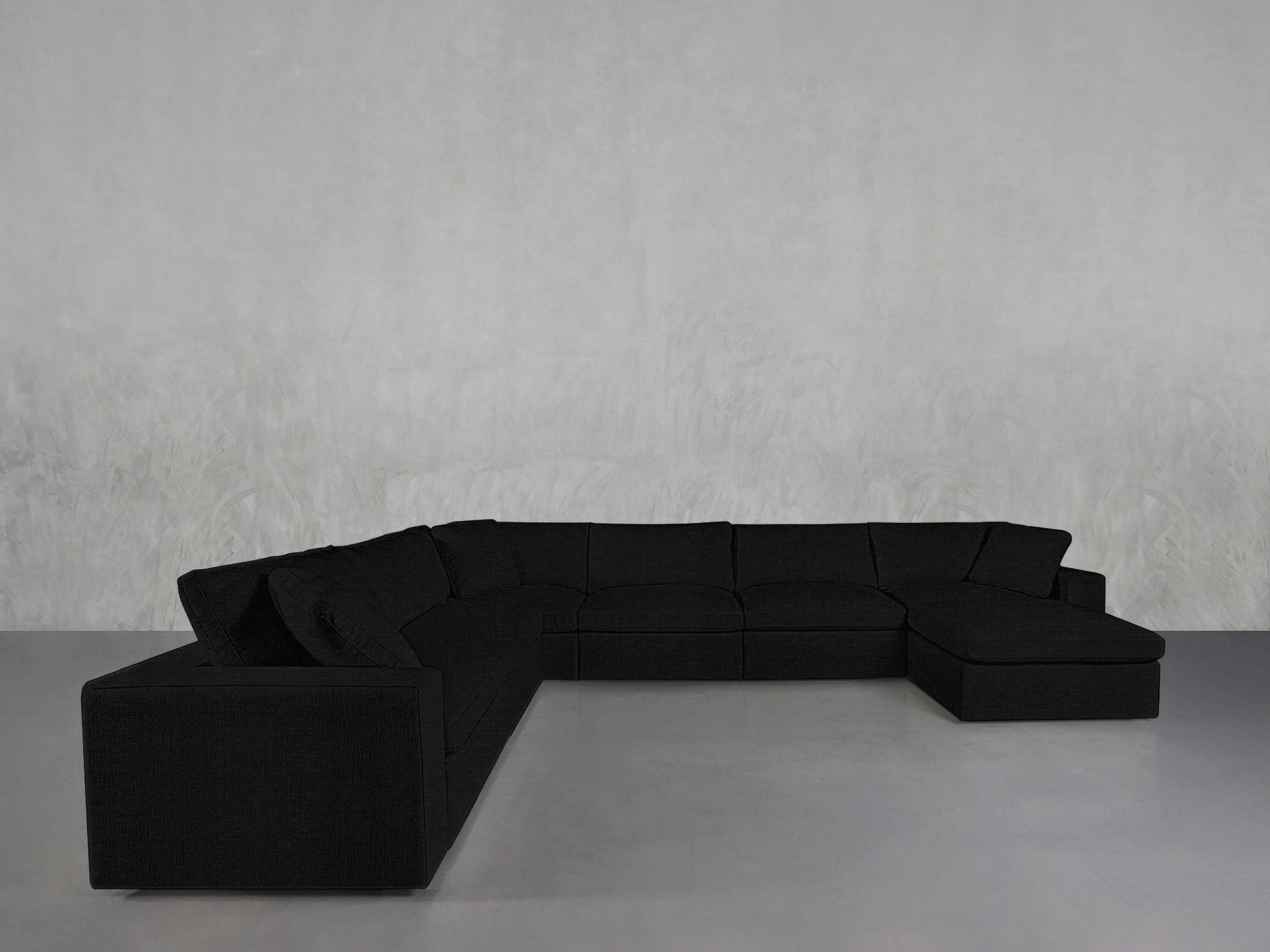 A 7-Seat Modular Chaise Corner Sectional in black, featuring clean lines and an L-shape, is set against a light gray textured wall on a smooth, light gray floor.