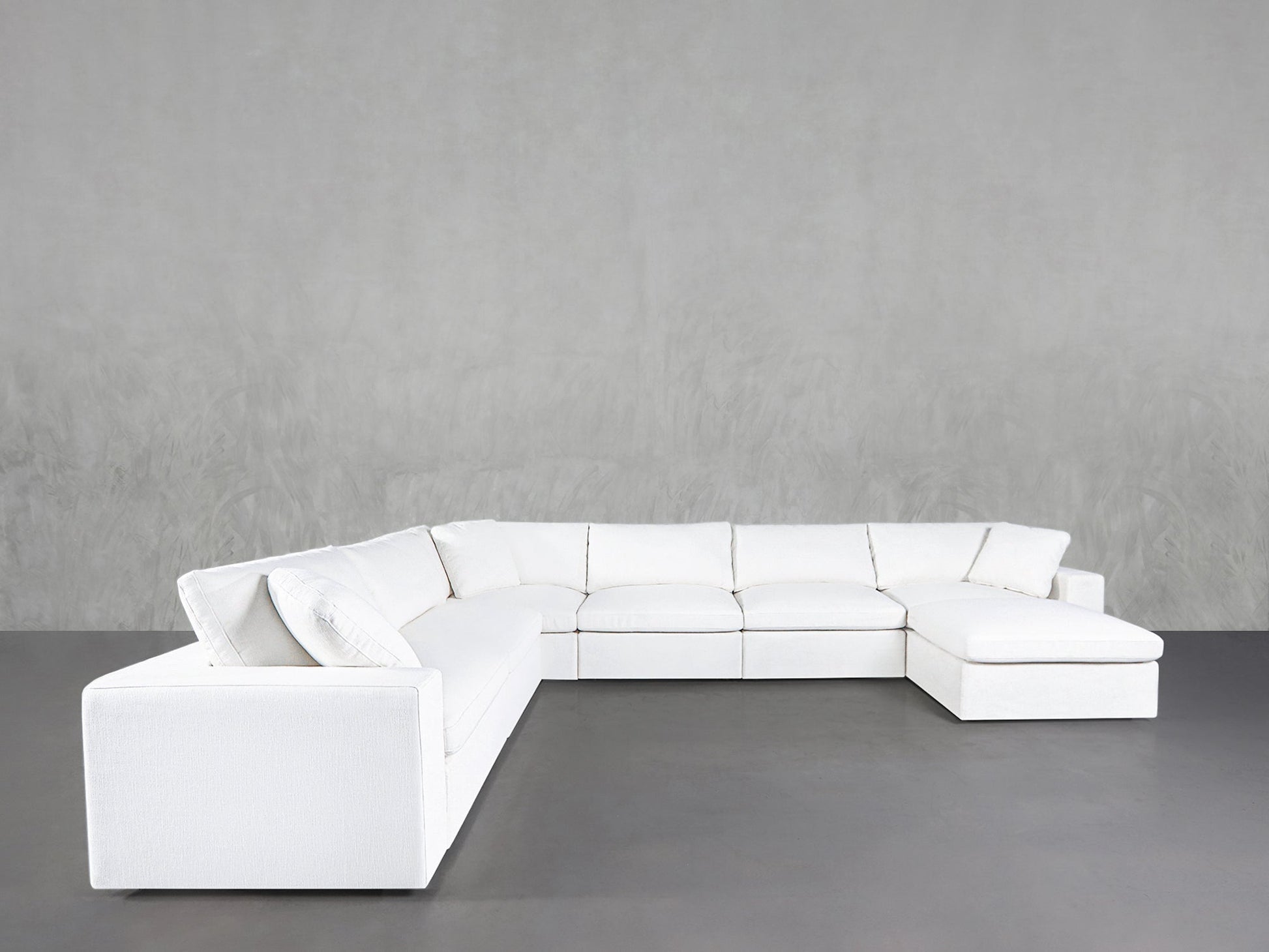 The 7-Seat Modular Chaise Corner Sectional, featuring clean lines and plush cushions, is arranged in an L-shape against a plain light gray wall and dark gray floor.