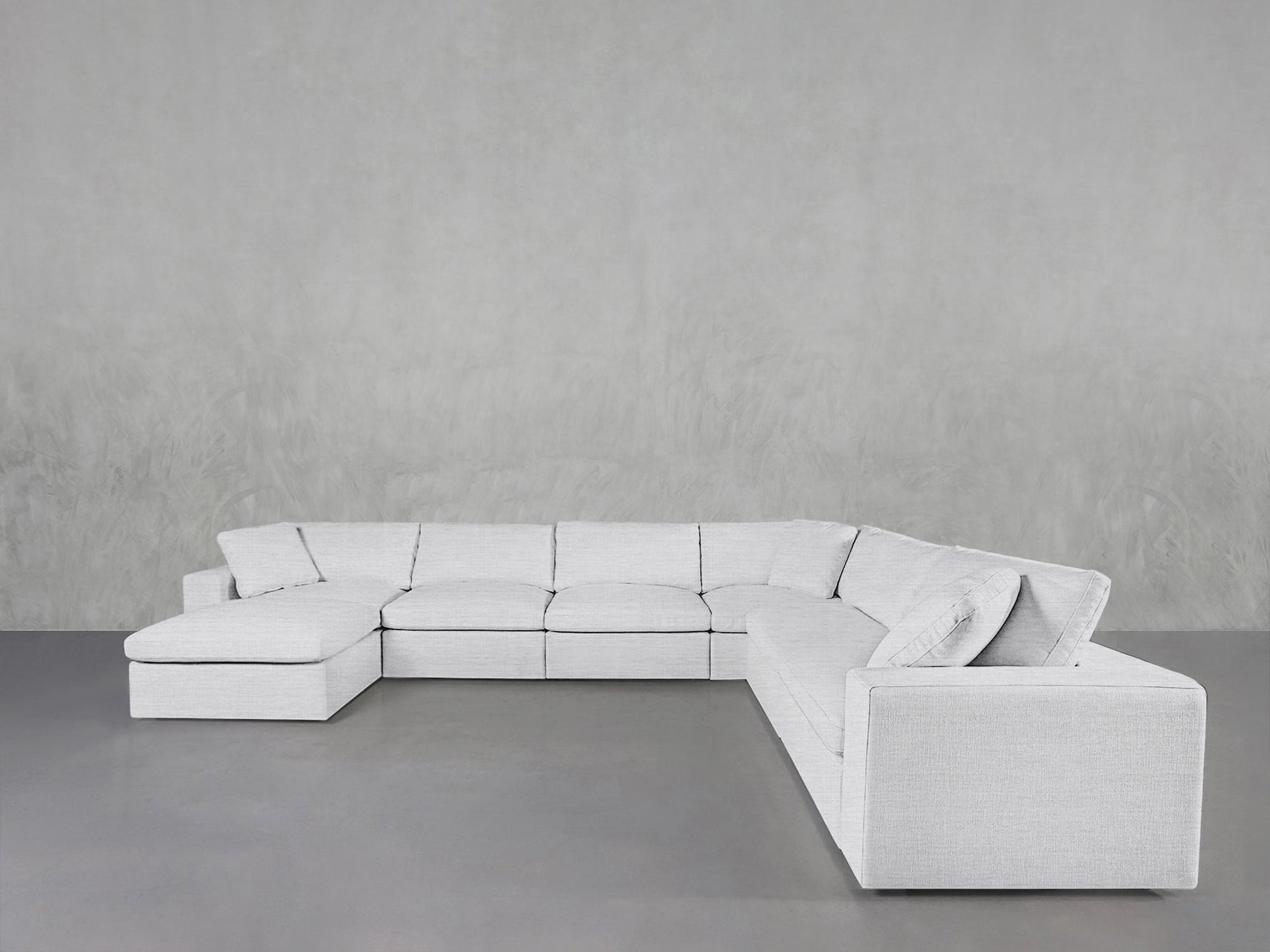 A 7-Seat Modular Chaise Corner Sectional in modern light gray features low backrests and square armrests, set against a plain light gray textured wall and a smooth gray floor.