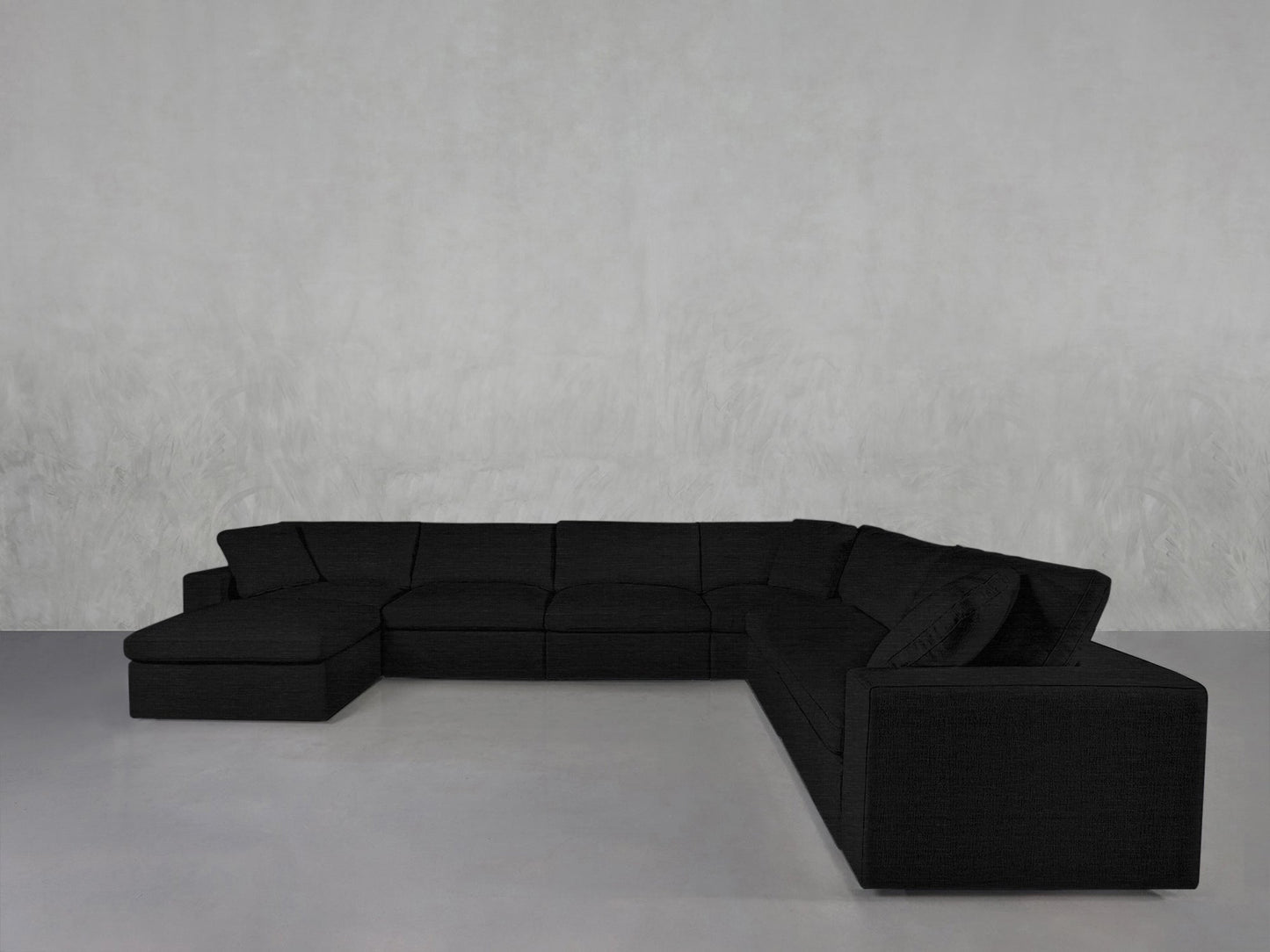 A 7-Seat Modular Chaise Corner Sectional in dark gray with a left-side chaise sits against a plain, light gray wall on a smooth, light-colored floor.
