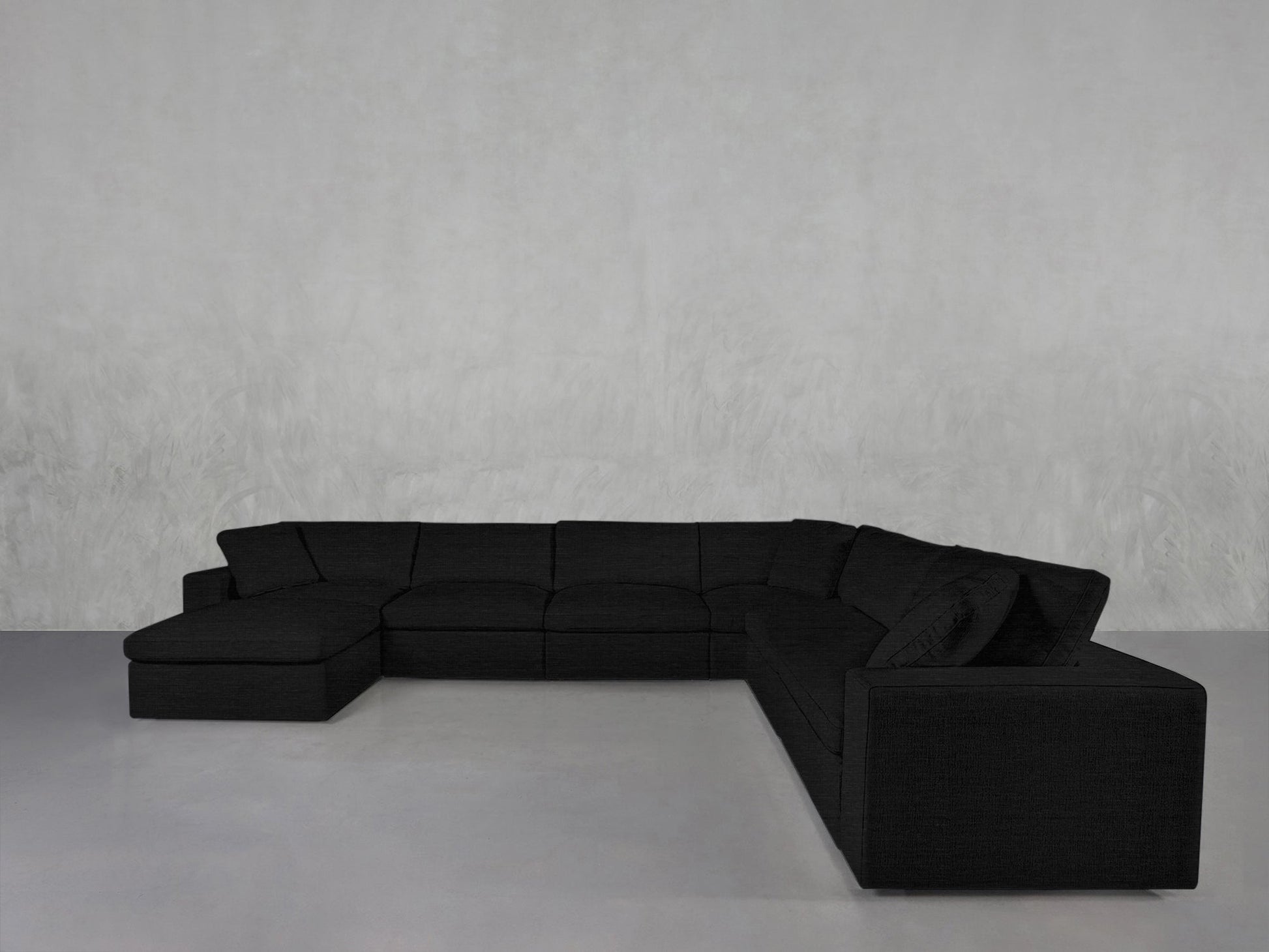 A 7-Seat Modular Chaise Corner Sectional in dark gray with a left-side chaise sits against a plain, light gray wall on a smooth, light-colored floor.