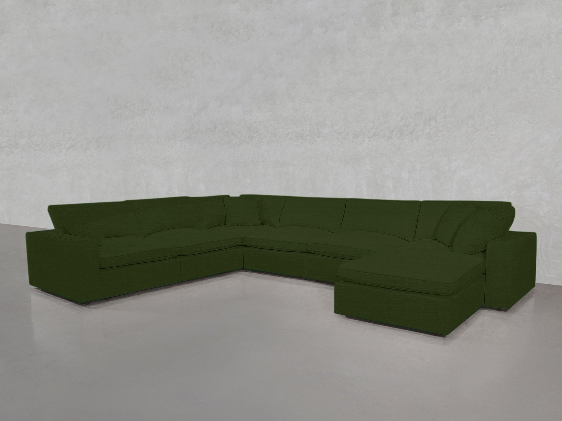 A spacious 7-Seat Modular Chaise Corner Sectional in dark green, featuring plush cushions, sits in a minimal light gray room with smooth floors and a textured wall.