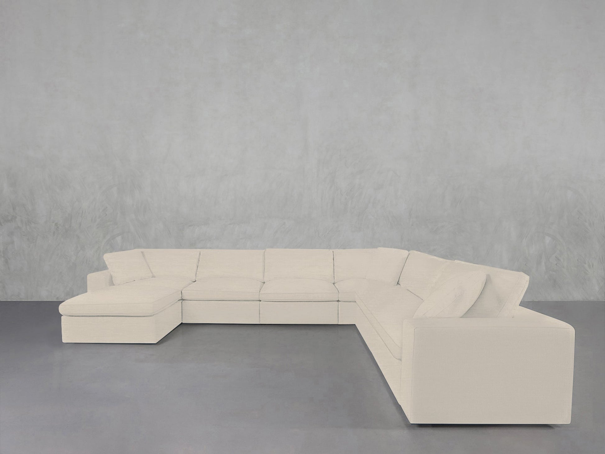 A 7-Seat Modular Chaise Corner Sectional in white sits on a gray floor against a light gray textured wall, creating a minimalist and spacious atmosphere.