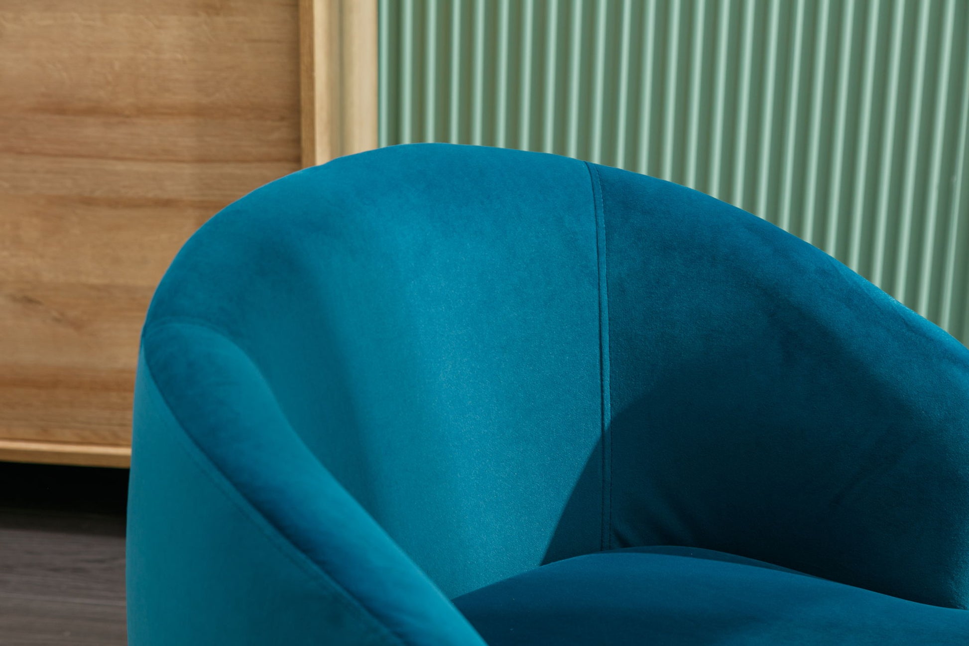 A close-up of the Velvet Fabric Swivel Accent Armchair Barrel Chair With Powder Coating Metal Ring in teal, featuring curved lines, set before a light green, vertically grooved wall and a wooden cabinet.