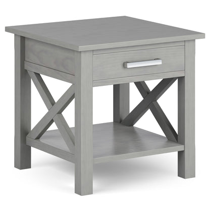 The Kitchener - Handcrafted Table is a gray wooden end table with a single drawer, metal handle, X-shaped side supports, and a lower open shelf.